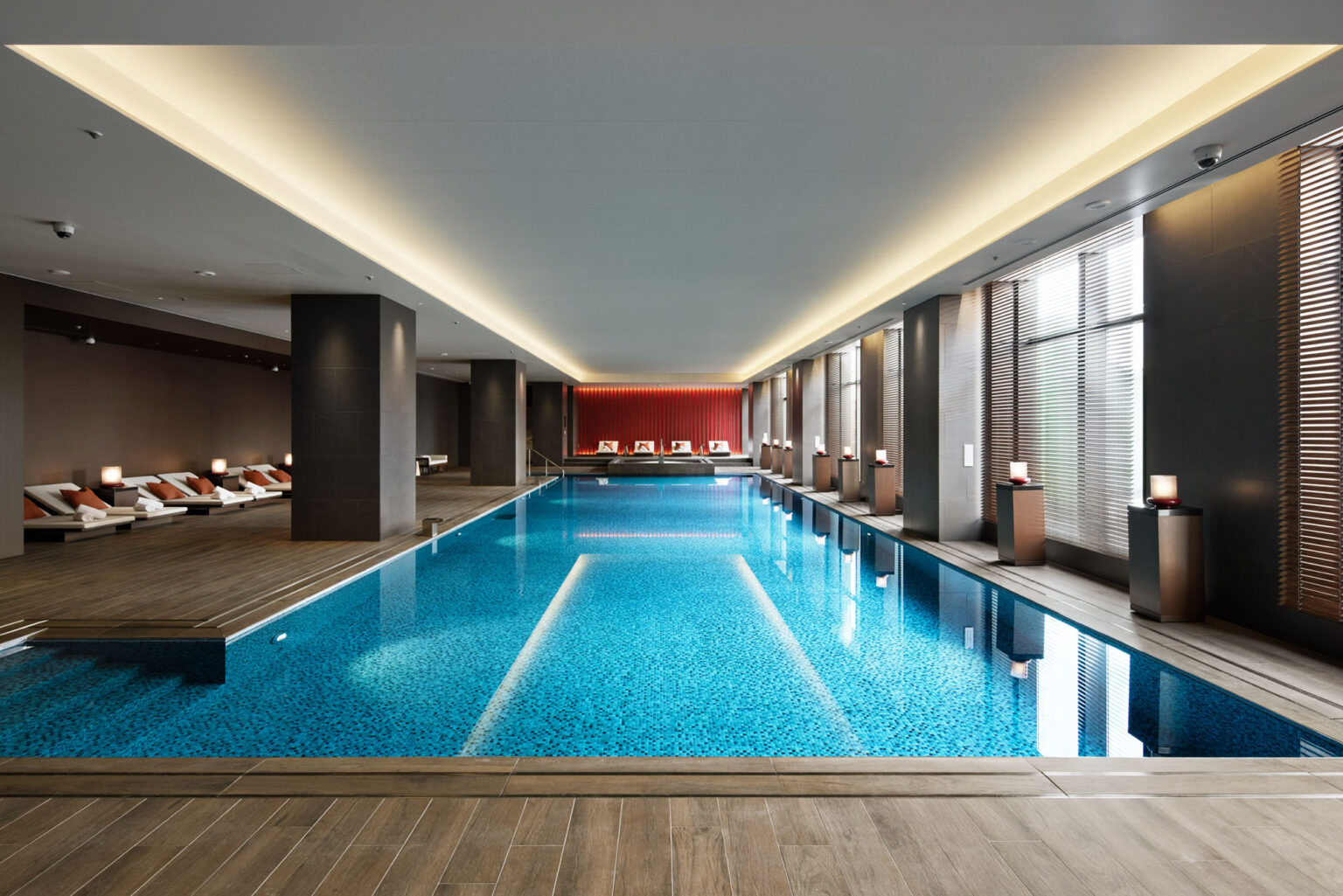 Luxurious indoor swimming pool of a hotel with clear blue water, surrounded by elegant loungers and soft lighting. Large windows on the right let in natural light.