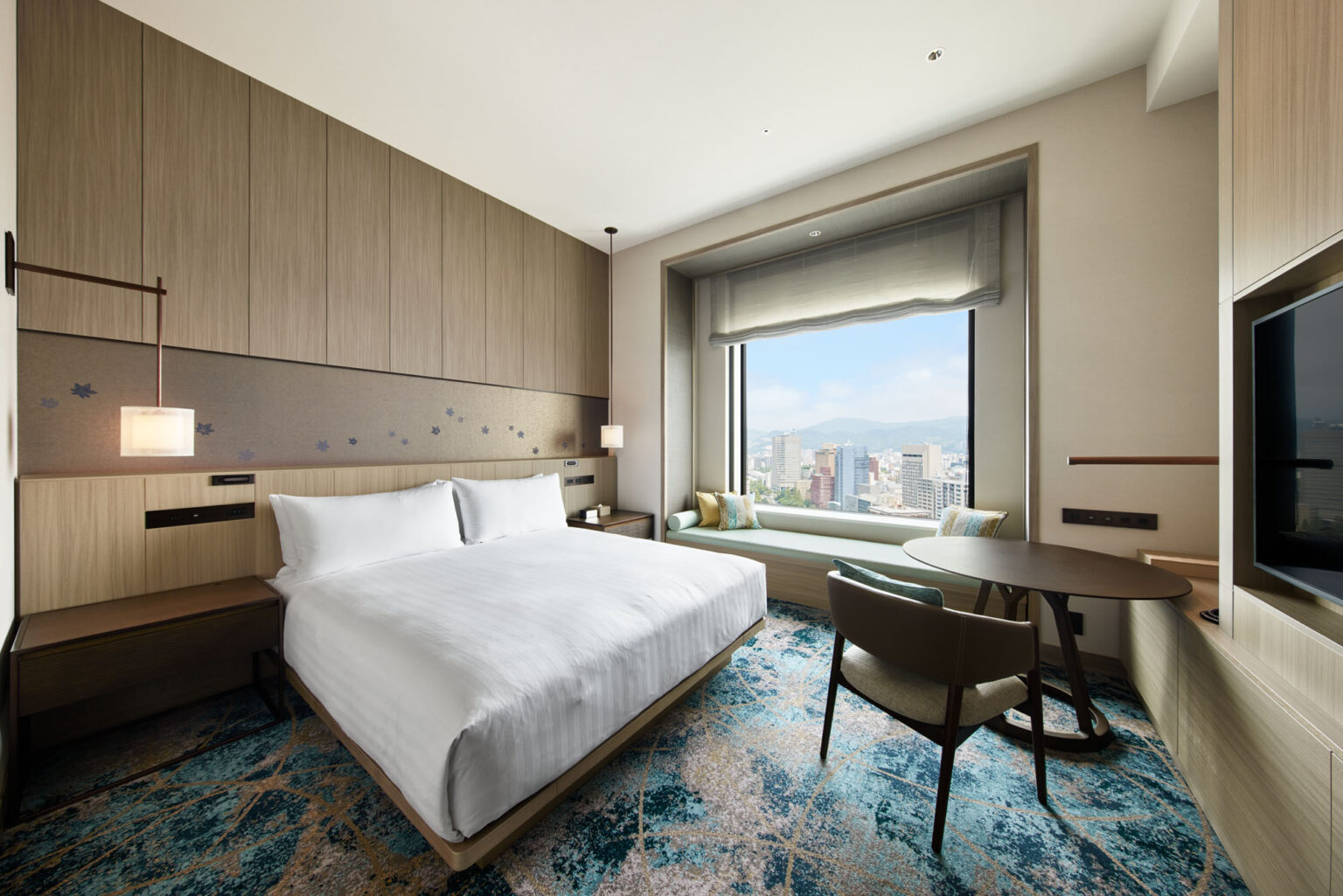 Modern hotel room featuring a large bed with white linens, a stylish chair at a round table, and a scenic city view through a big window. Calm, inviting atmosphere.