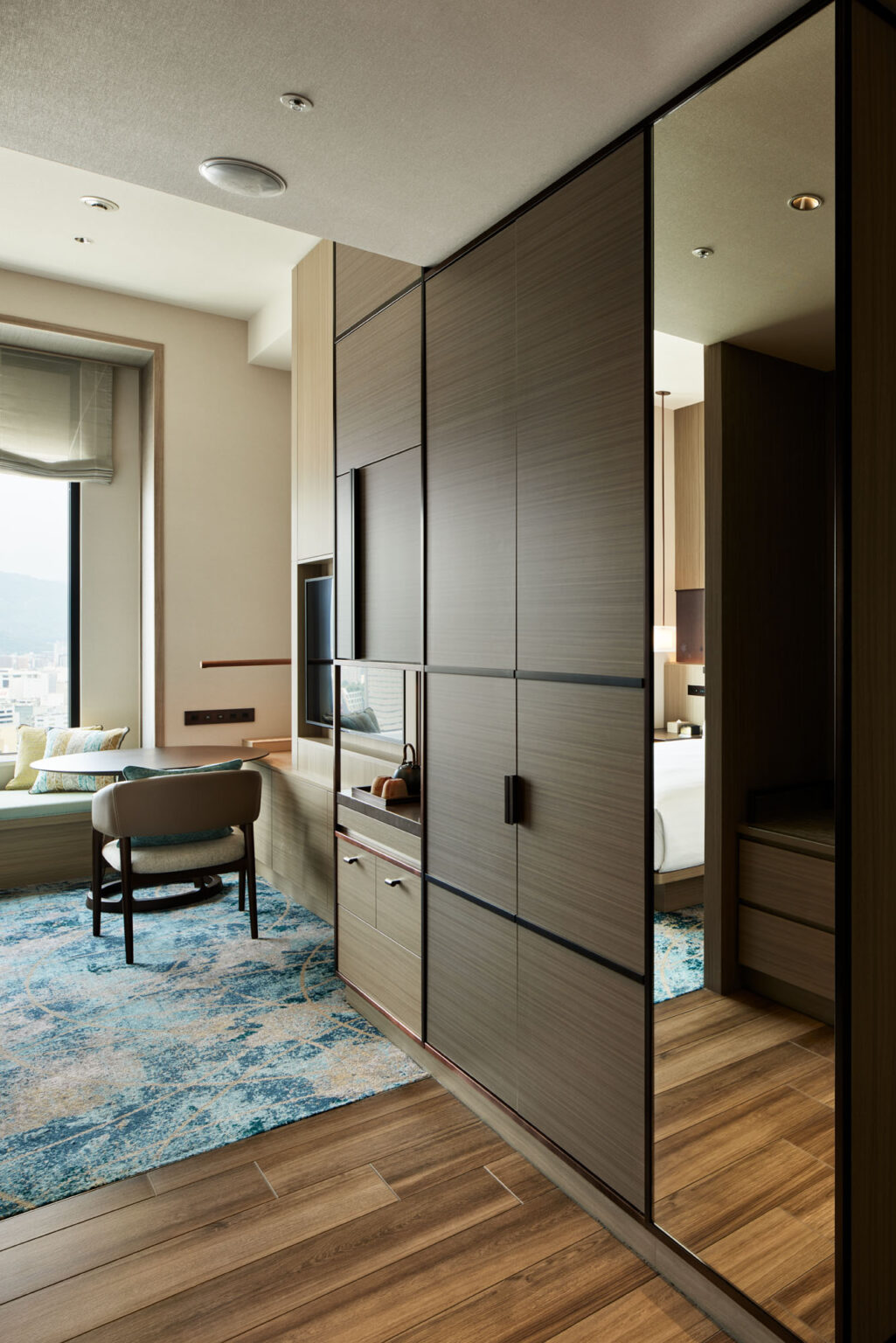 Modern hotel room with wooden accents, a large window, a blue patterned carpet, and a cozy sitting area. The space feels serene and inviting.