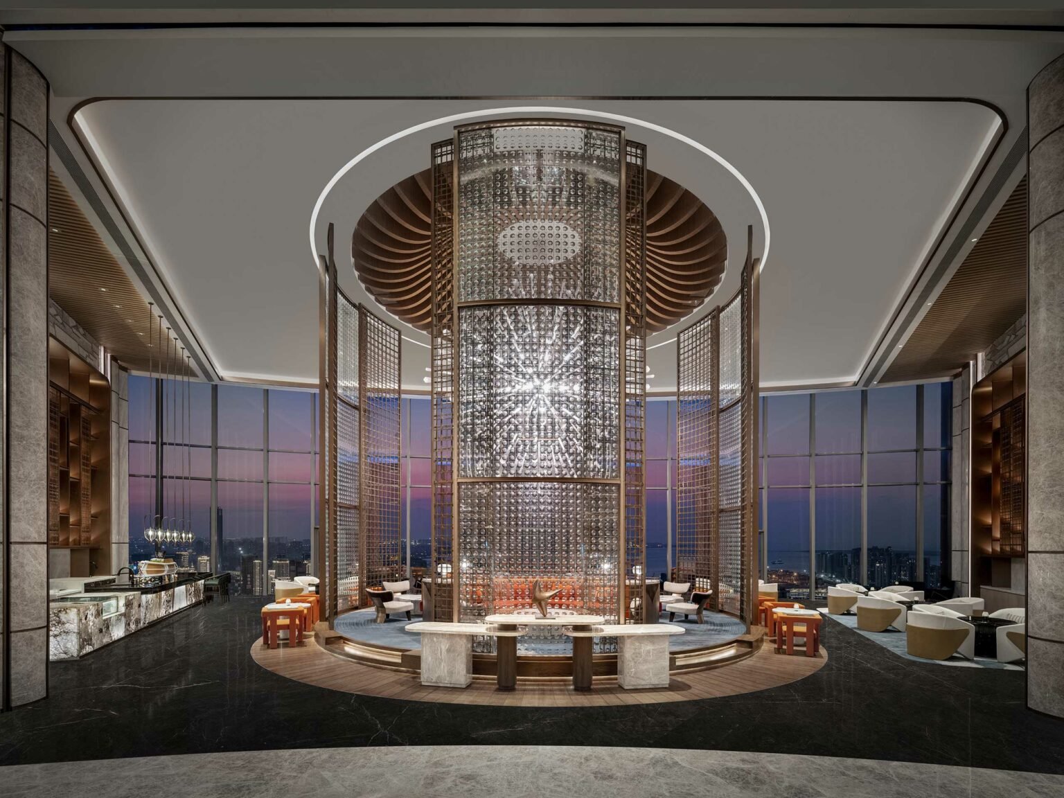 Luxurious modern hotel lounge with sleek decor featuring a central cylindrical light fixture, surrounded by elegant seating. The floor-to-ceiling windows reveal a stunning cityscape at dusk.
