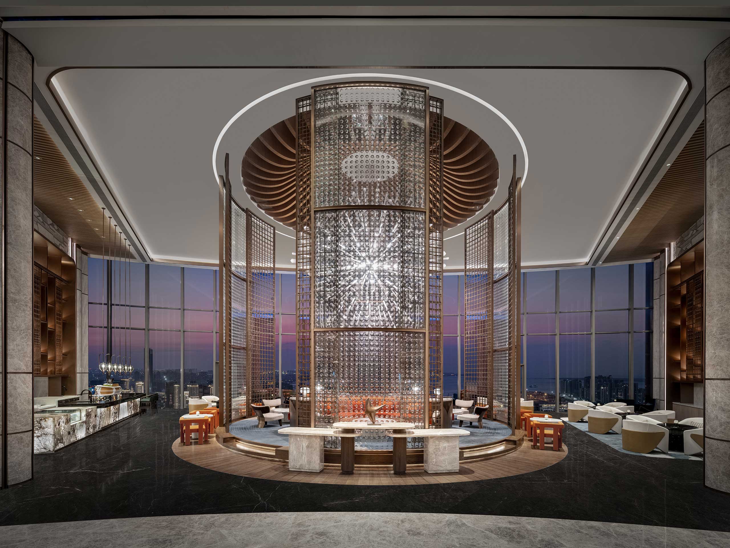 Luxurious modern hotel lounge with sleek decor featuring a central cylindrical light fixture, surrounded by elegant seating. The floor-to-ceiling windows reveal a stunning cityscape at dusk.