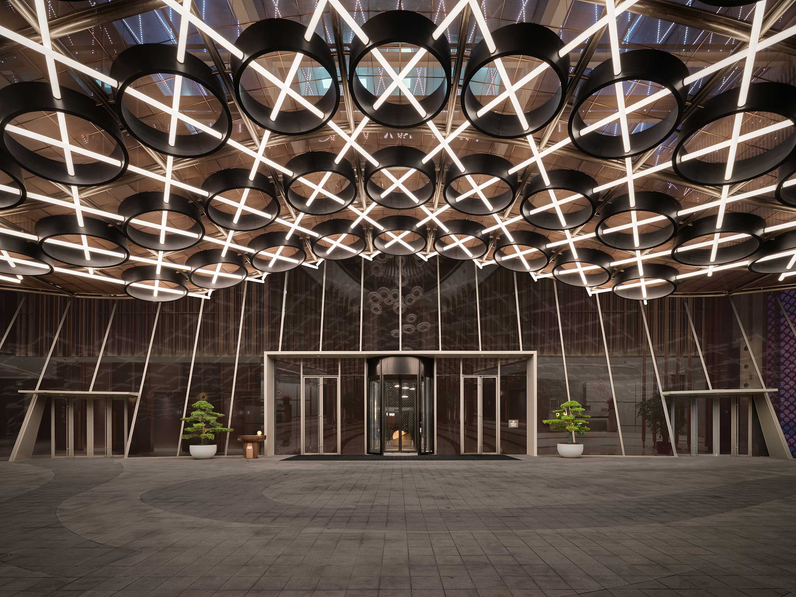 Modern hotel lobby entrance with circular, cross-patterned ceiling lights. Glass doors are centered, flanked by potted plants. Bright, sleek, and symmetric design.