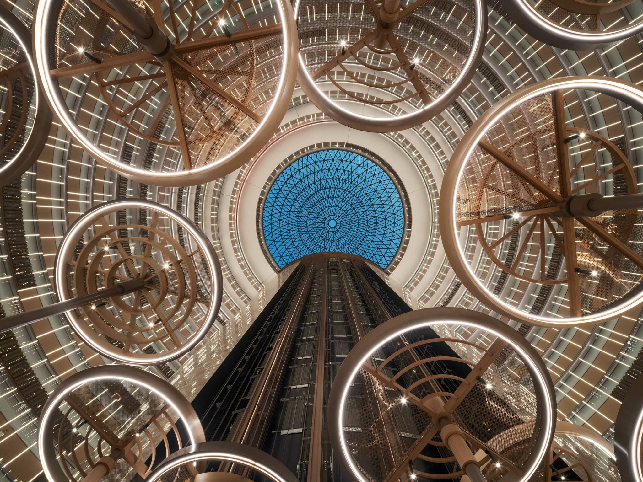 View up an expansive hotel atrium with glowing circular light fixtures and a blue glass dome ceiling, creating a futuristic and elegant atmosphere.