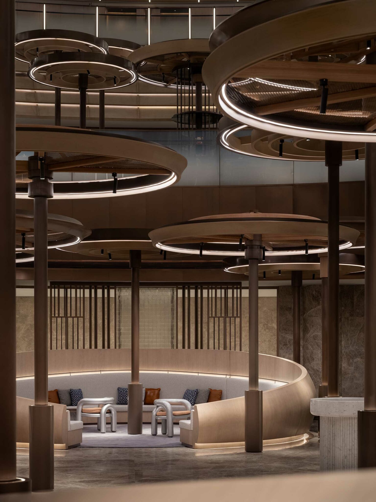 Modern hotel lounge with circular ceiling lights and brown columns above a cozy conversation pit featuring curved seating and cushioned chairs. Sophisticated ambiance.