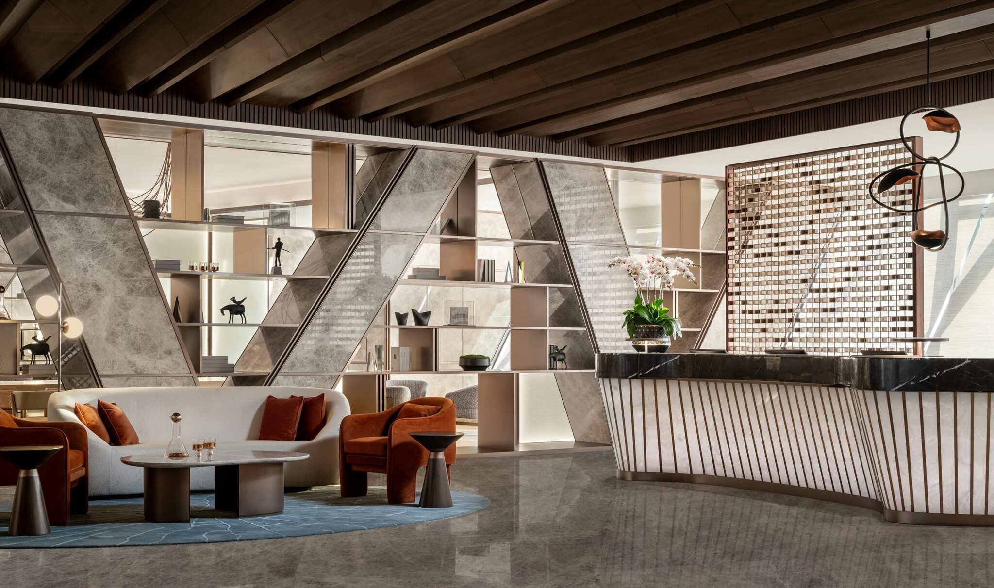 Modern hotel lobby with geometric decor. Features a sleek reception desk, orange armchairs, white sofa, shelves, and a plant. Warm, welcoming atmosphere.