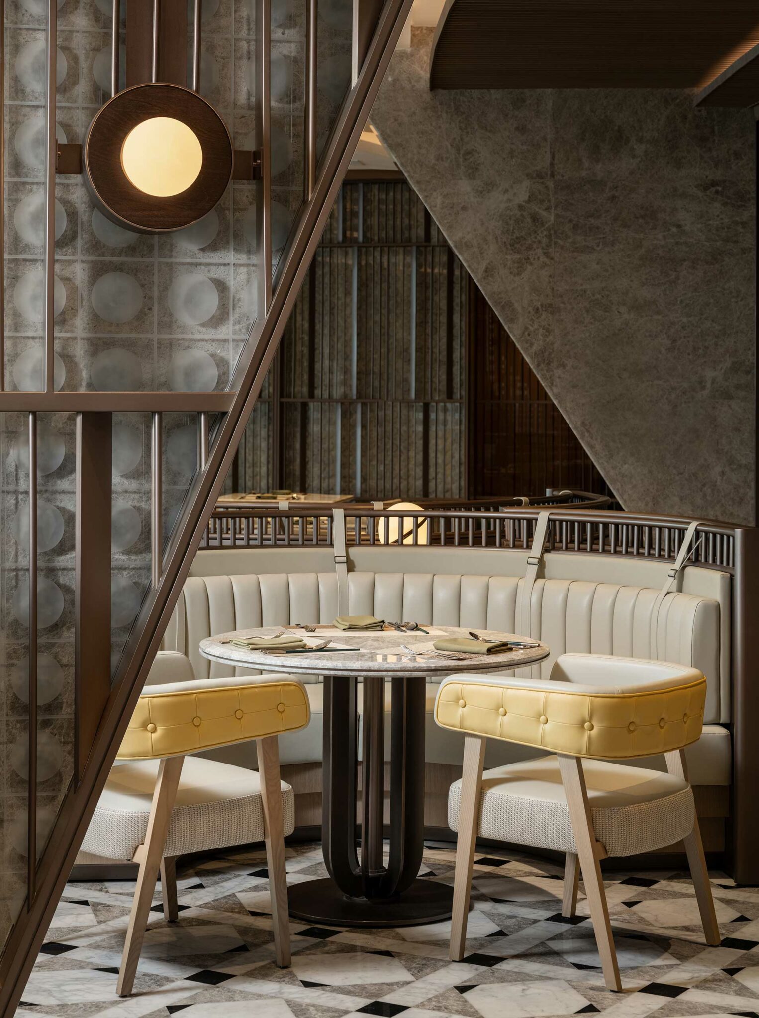 A modern hotel restaurant interior with a round table, yellow chairs, and a beige cushioned booth. Geometric patterns and warm lighting create an elegant atmosphere.