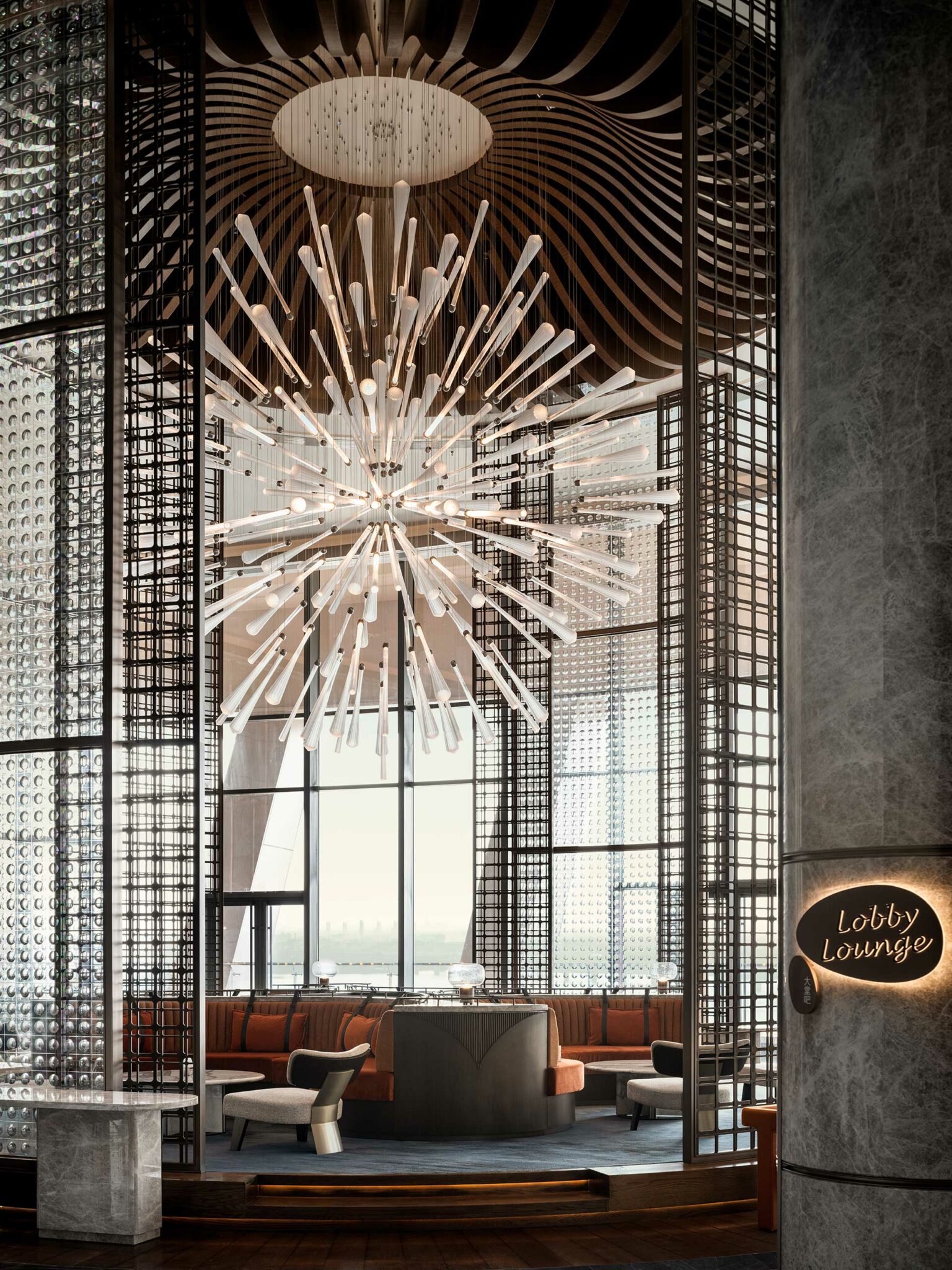 Modern hotel lounge with a starburst chandelier above a circular seating area. Tall windows and textured walls create a sophisticated atmosphere.