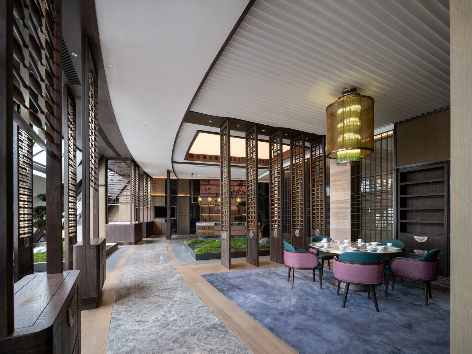 Spacious, modern hotel interior with curved ceiling, wooden accents, and large windows. A dining area features a round table with colorful chairs, exuding elegance.