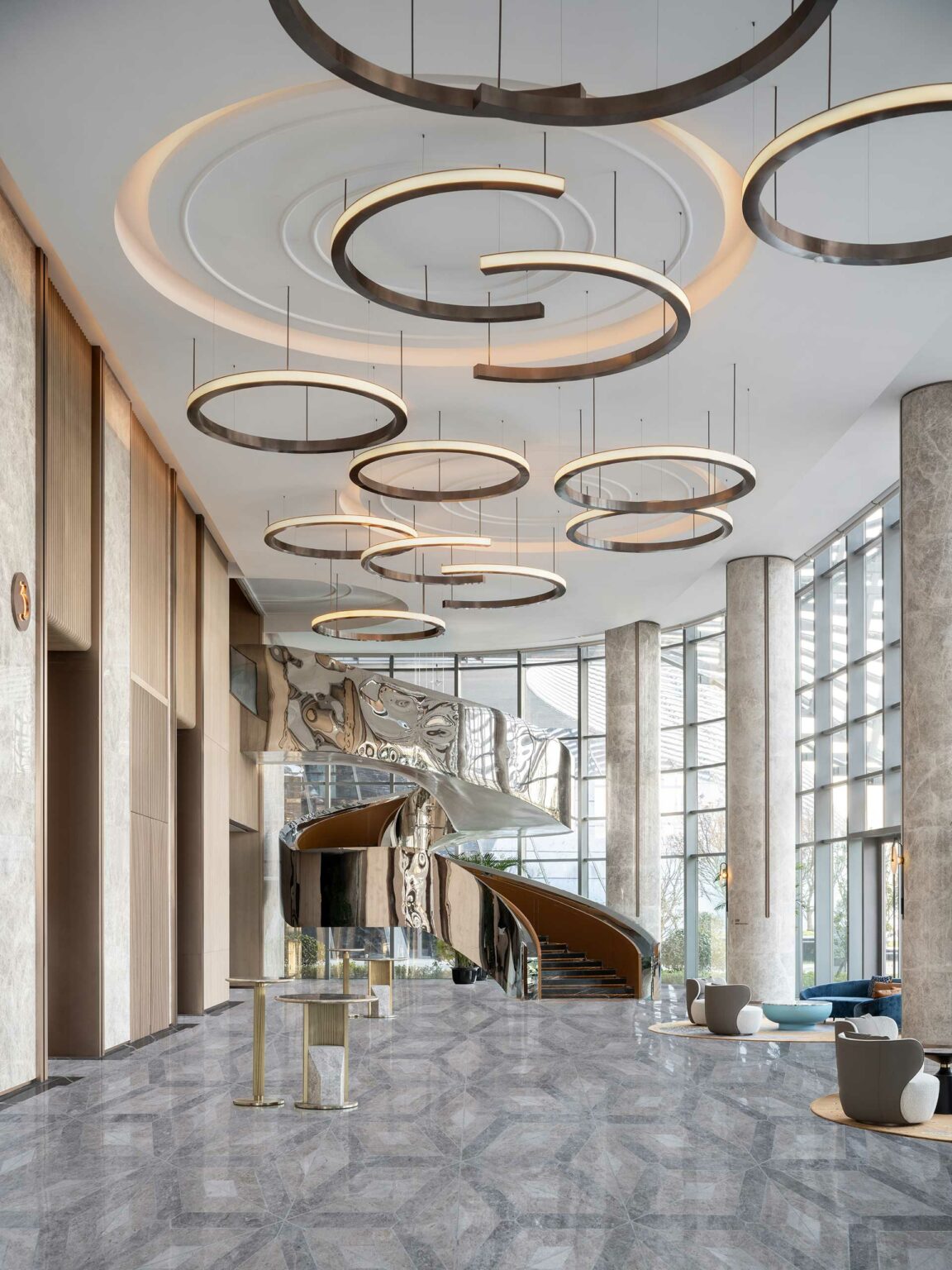 Spacious modern hotel lobby with elegant ring chandeliers, tall windows, and a curving metal staircase. The tone is sleek and sophisticated.