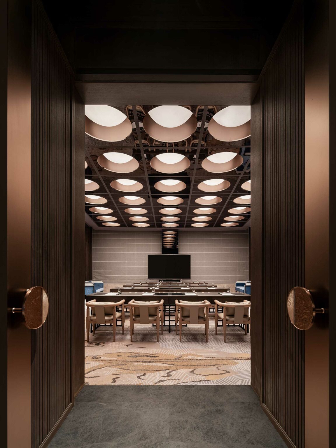 Open doors reveal a hotel conference room with modern decor. Circular ceiling lights, a central TV, and cushioned chairs evoke a sleek, professional tone.