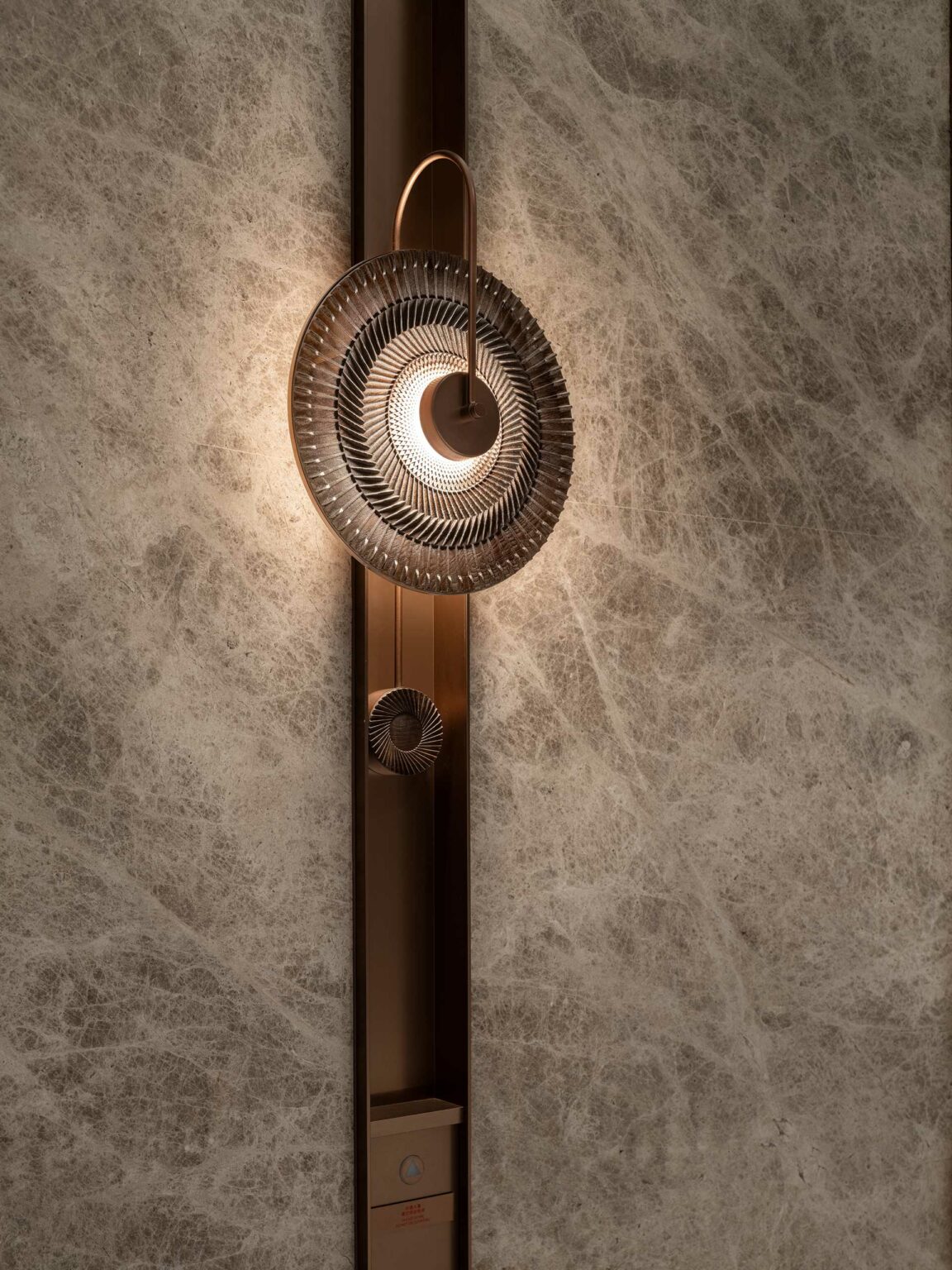Elegant elevator wall sconce with circular, spiral design casting warm light. Mounted on textured beige wall, creating a sophisticated ambiance.