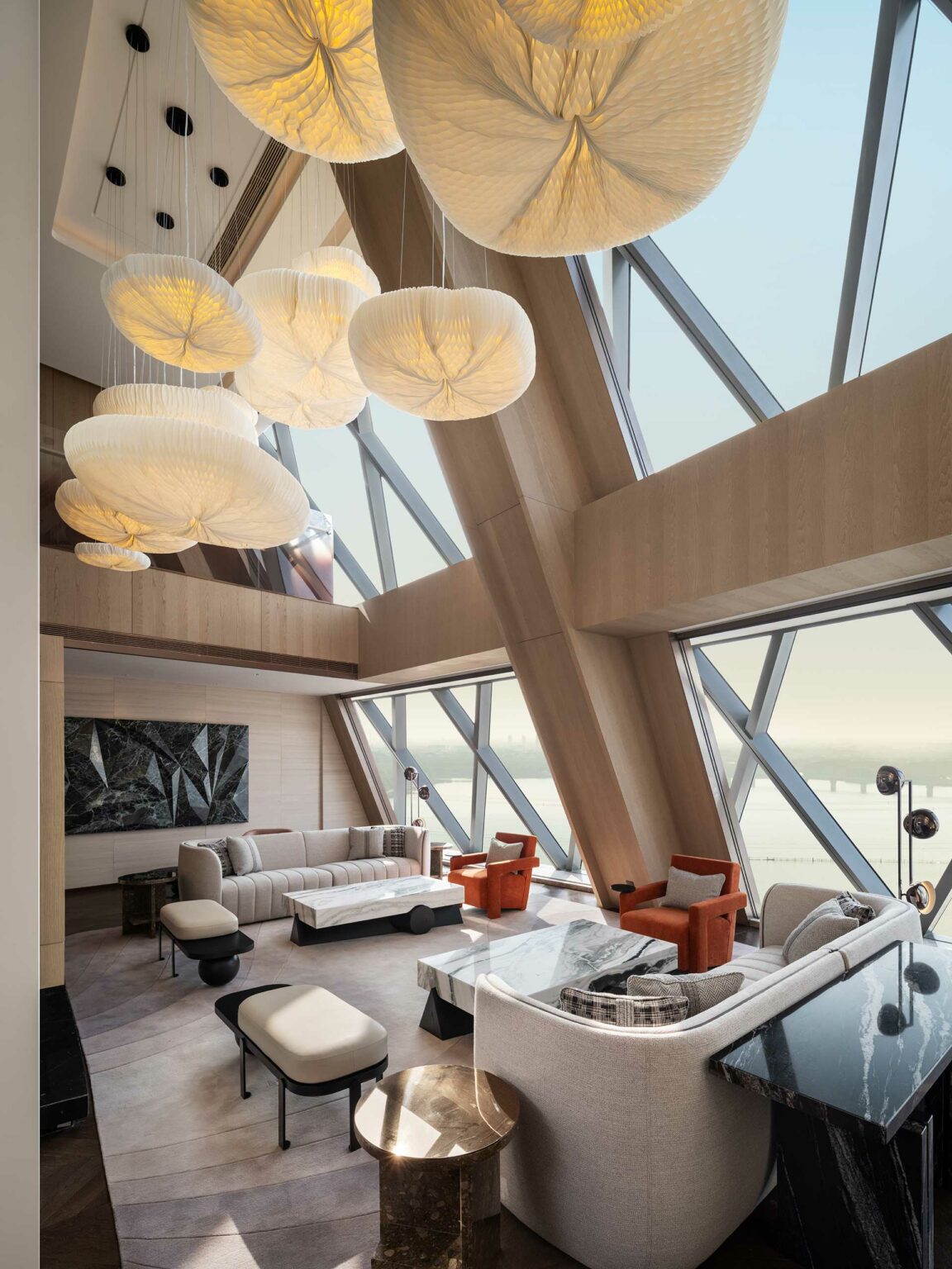 A modern suite living room with large geometric windows and soft lighting. Features include abstract pendant lights, plush sofas, and orange armchairs, creating a cozy yet elegant atmosphere.