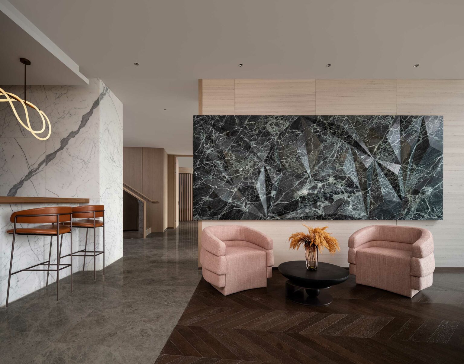 Modern hotel lounge features two pink armchairs, round black table with dry floral arrangement, large marble wall art, and a marble bar with stools.