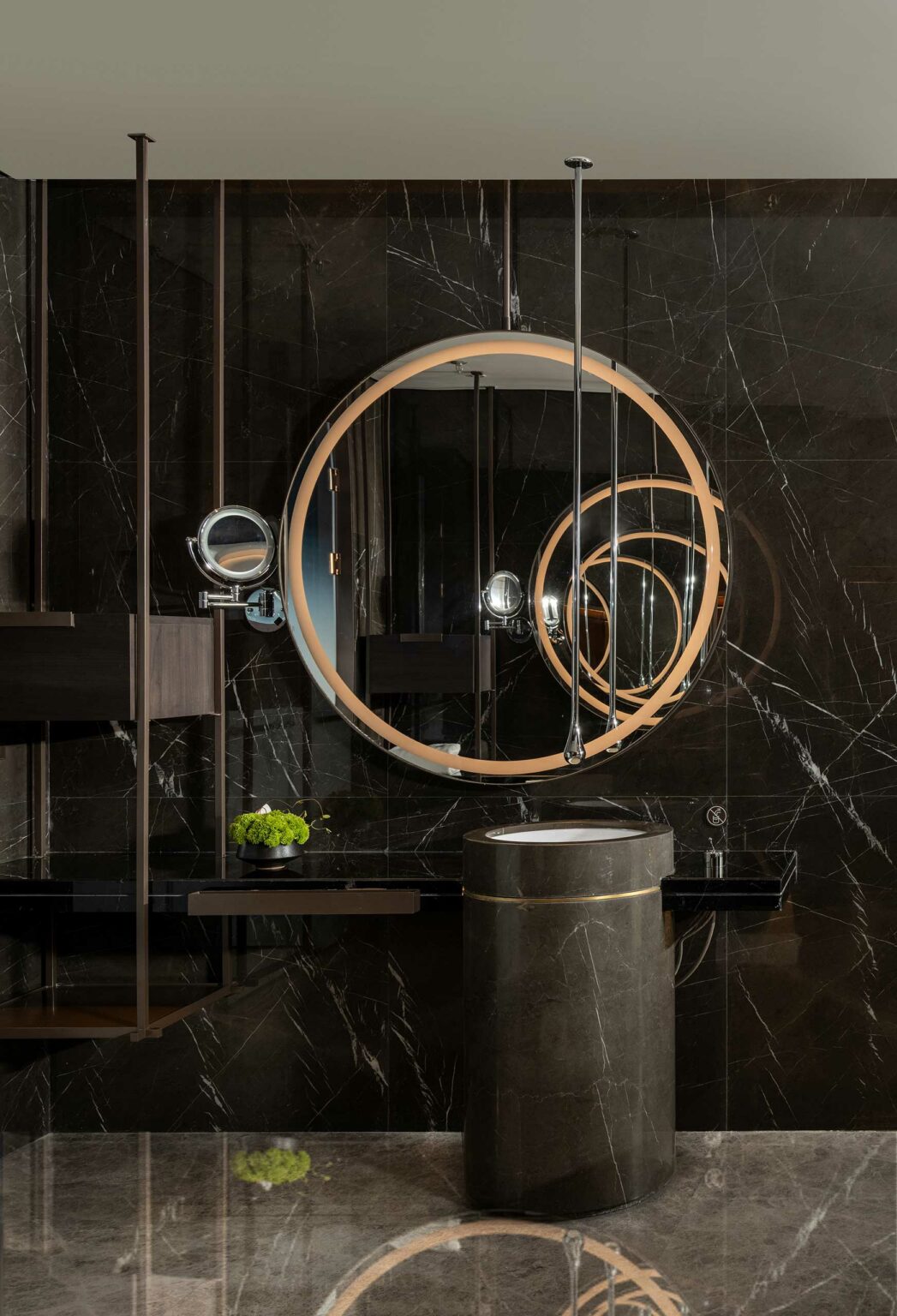 Luxurious suite bathroom with a large round mirror on a dark marble wall, reflecting a modern, sleek decor. A cylindrical sink stands below, with small greenery adding a fresh touch.