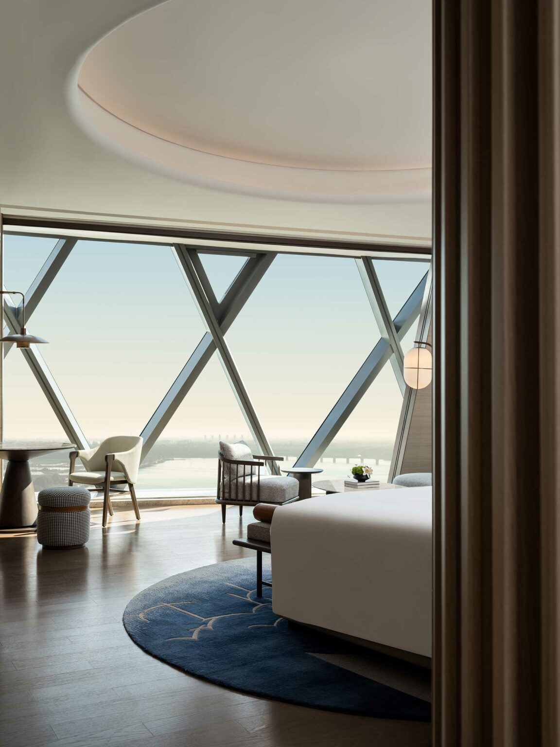 Modern hotel room with large triangular windows offering a scenic view. Features a cozy bed, stylish chairs, circular rug, and a calm, inviting ambiance.