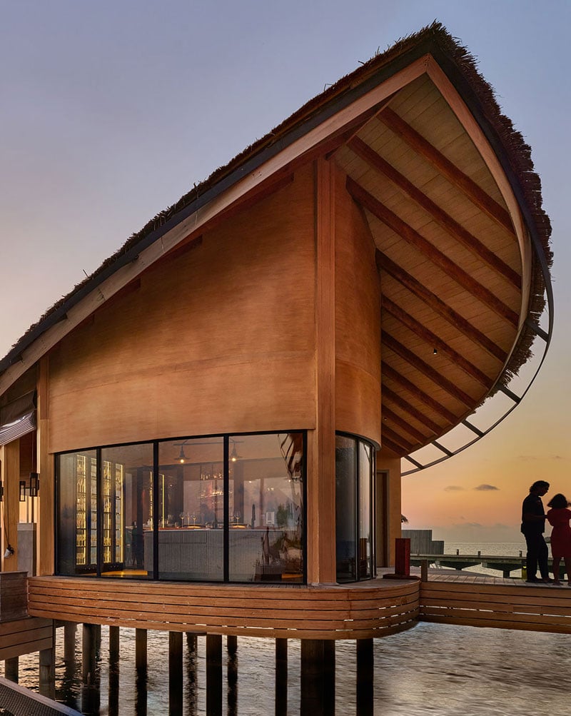 Curved wooden structure on stilts over water at sunset, featuring large glass windows. A person stands silhouetted on a deck, creating a serene mood.