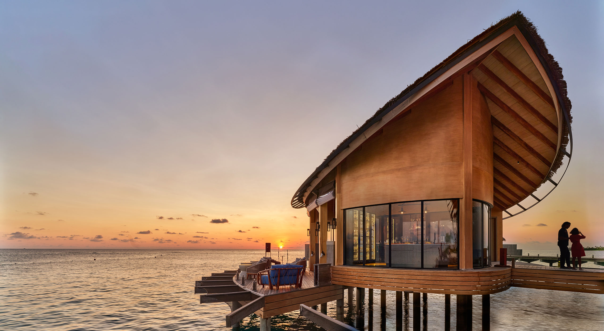 Modern overwater resort bungalow with wooden architecture extending into the ocean at sunset. Warm hues reflect on the water. Two silhouetted figures stand, adding tranquility.