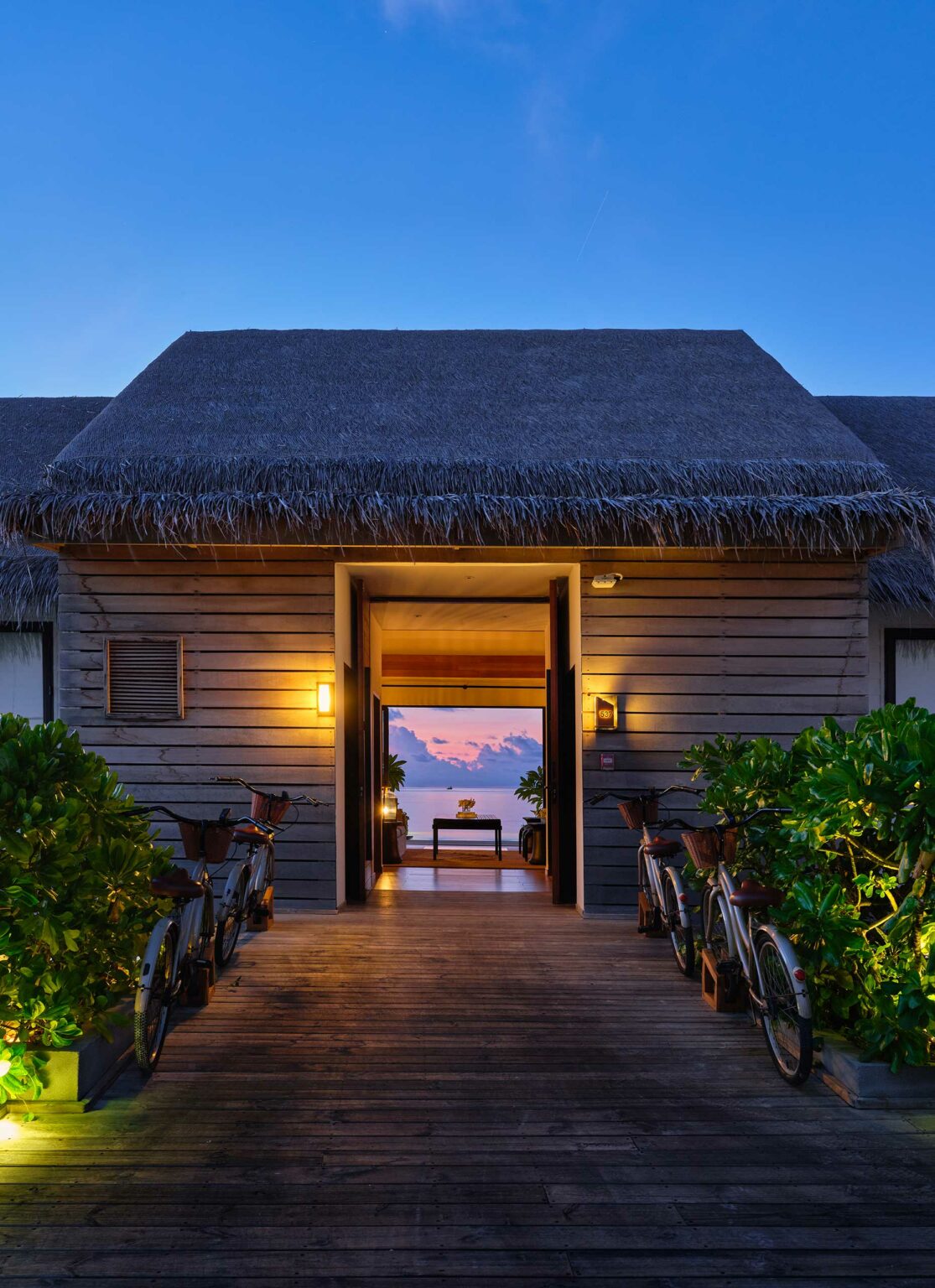 Wooden walkway leading to an open doorway of a thatched-roof resort bungalow. Bikes and lush greenery line the path. A sunset sky with clouds is visible inside.
