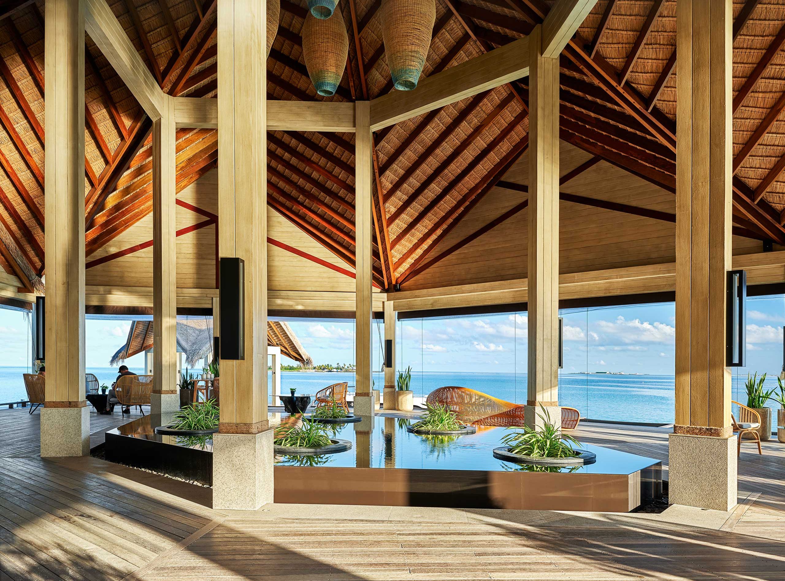 Spacious open-air resort lobby with high thatched roof, wooden beams, hanging wicker lights, and a reflecting pool. Ocean view creates a serene atmosphere.