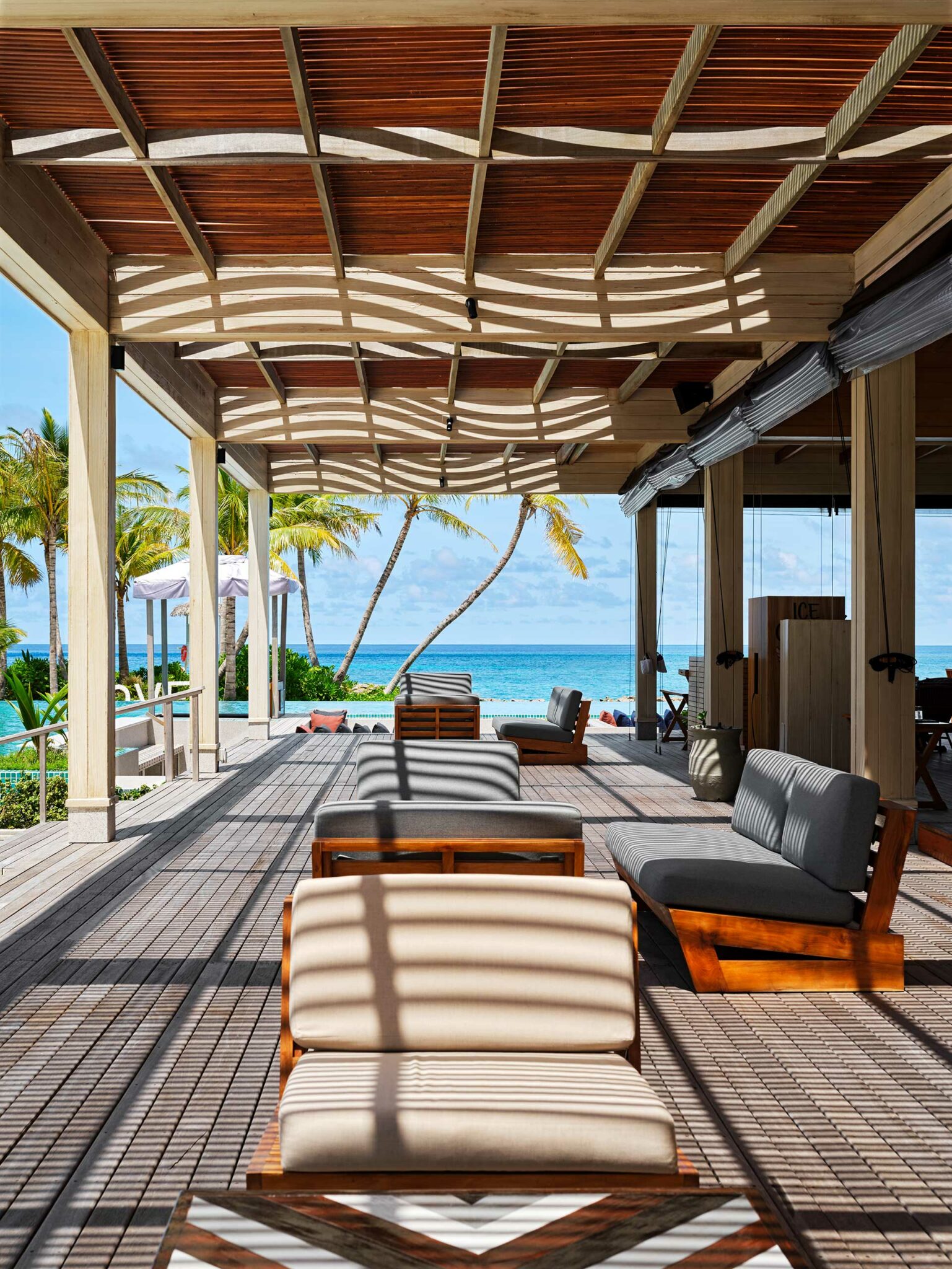 Open-air resort lounge with wooden seating under a sunlit, slatted roof. Palm trees and turquoise ocean are visible in the background, creating a serene atmosphere.