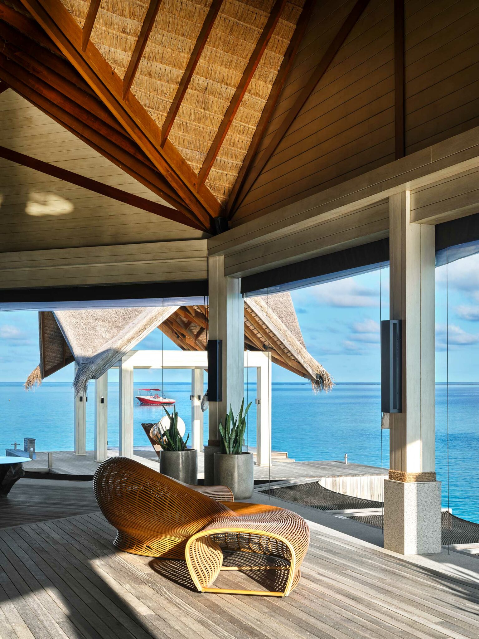 A stylish open-concept oceanfront resort room with a high thatched ceiling and wooden floors. A curved rattan chair overlooks a vibrant blue sea, creating a serene, tropical atmosphere.