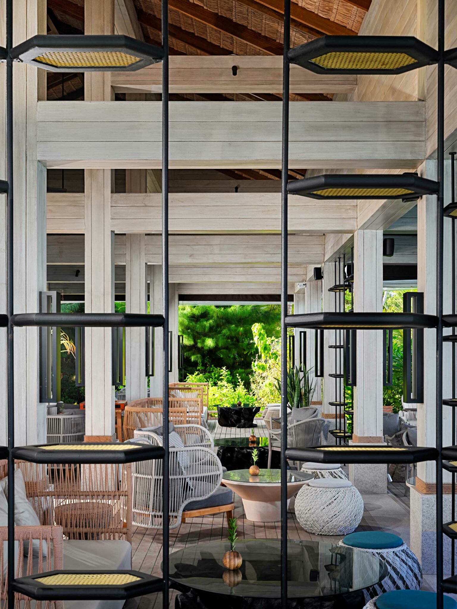 Open-air resort lounge with modern, white wood beams and wicker furniture. Glass tables and green plants create a serene, inviting atmosphere amidst lush greenery.