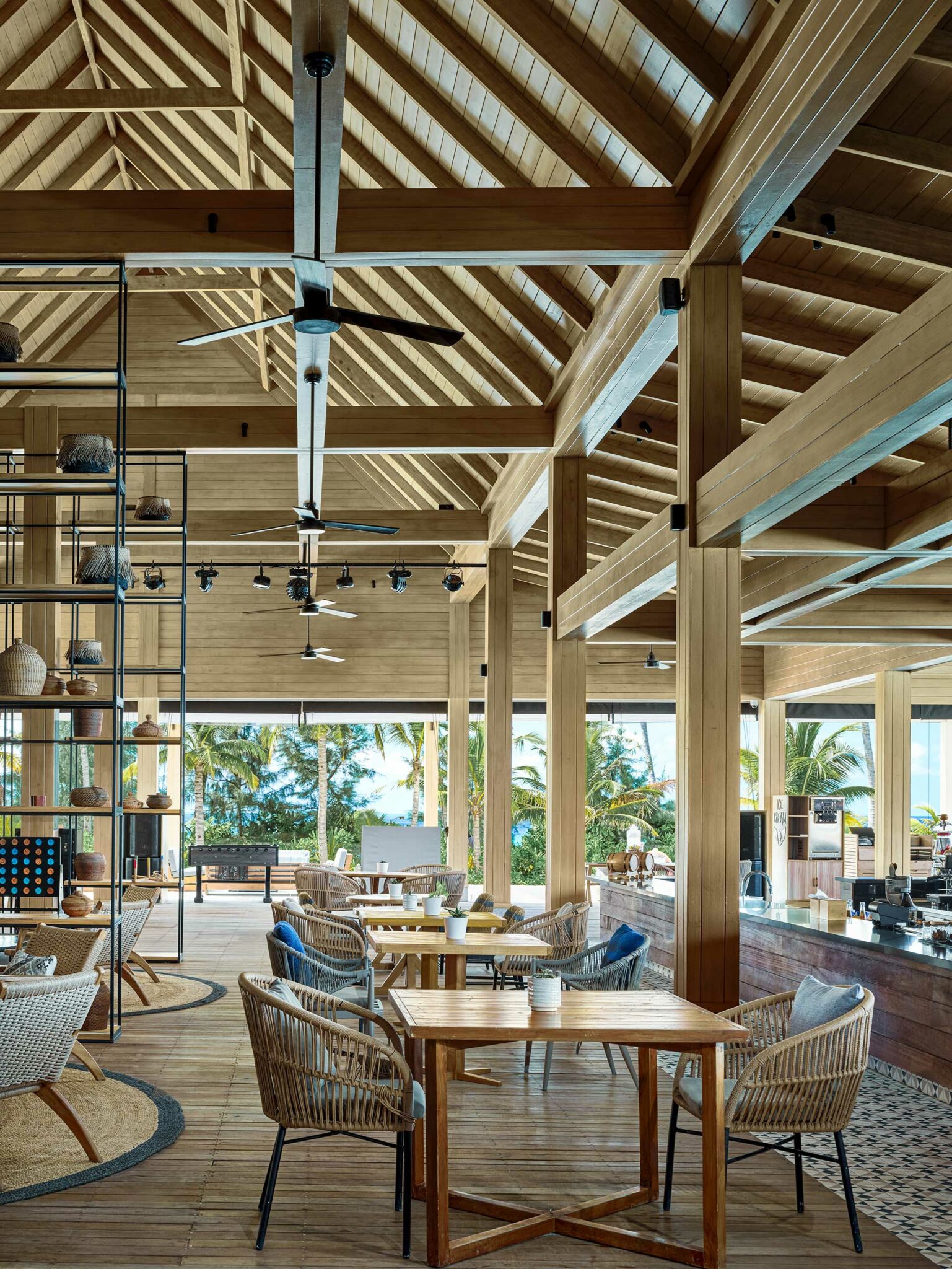 Open-air resort cafe with wooden beams and wicker chairs, featuring tables set for dining. Large windows reveal lush palm trees, creating a serene, tropical vibe.