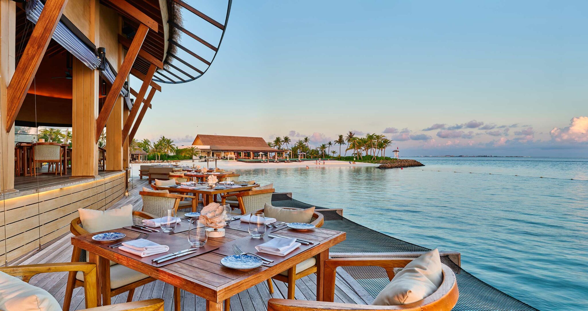 A serene seaside resort restaurant with wooden tables set for dining, overlooking calm blue waters at sunset. Palm trees in the distance under a clear sky.