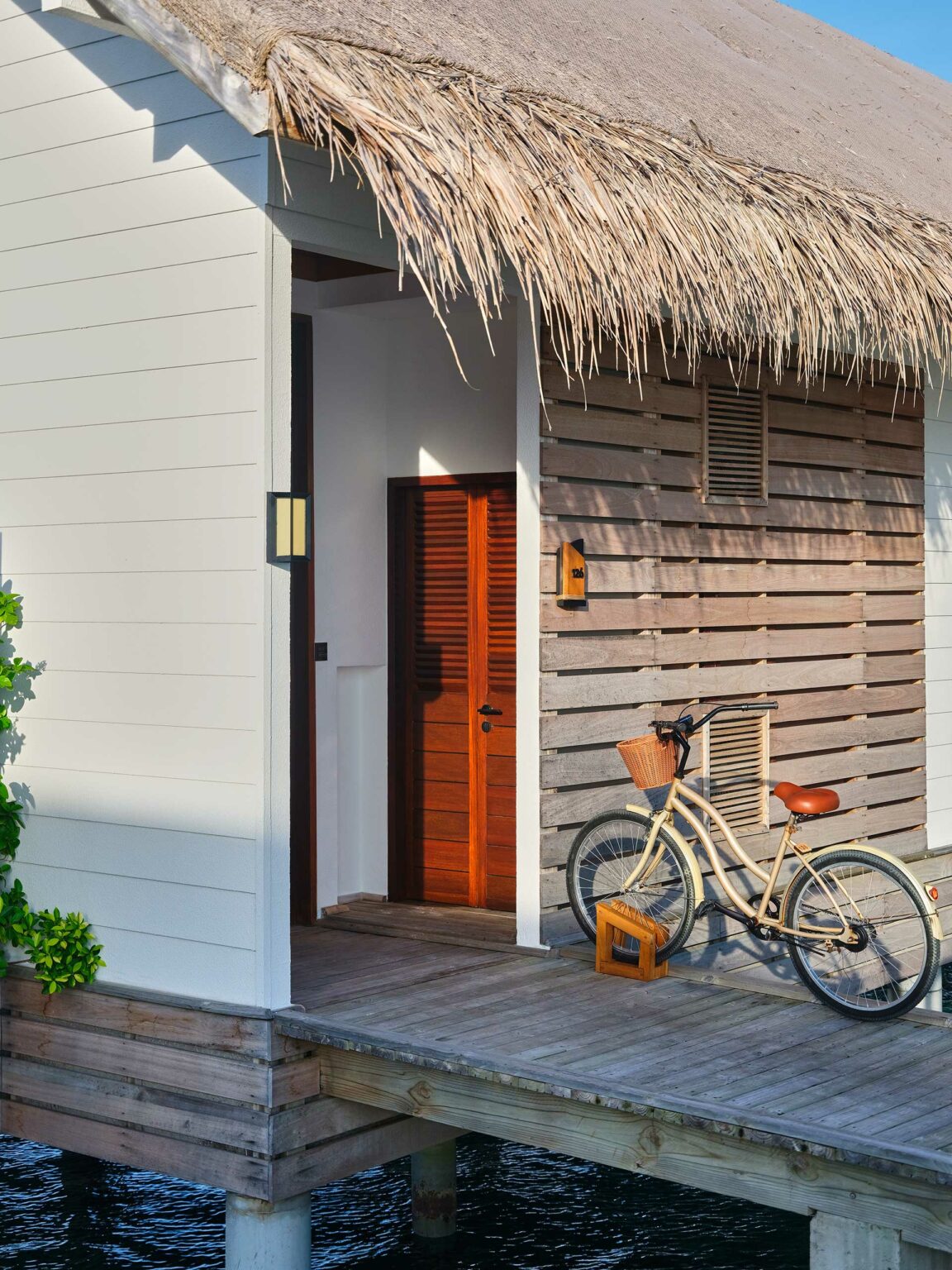 A cozy resort bungalow with a thatched roof and wooden walls stands on stilts. A beige bicycle with a wicker basket rests against the wall, conveying a tranquil, coastal vibe.