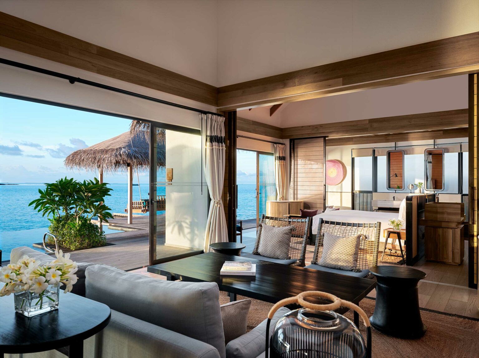 Luxurious open living space of a resort bungalow with light wood tones overlooks a serene ocean view. Large windows invite natural light. Cozy seating with tropical decor enhances tranquility.