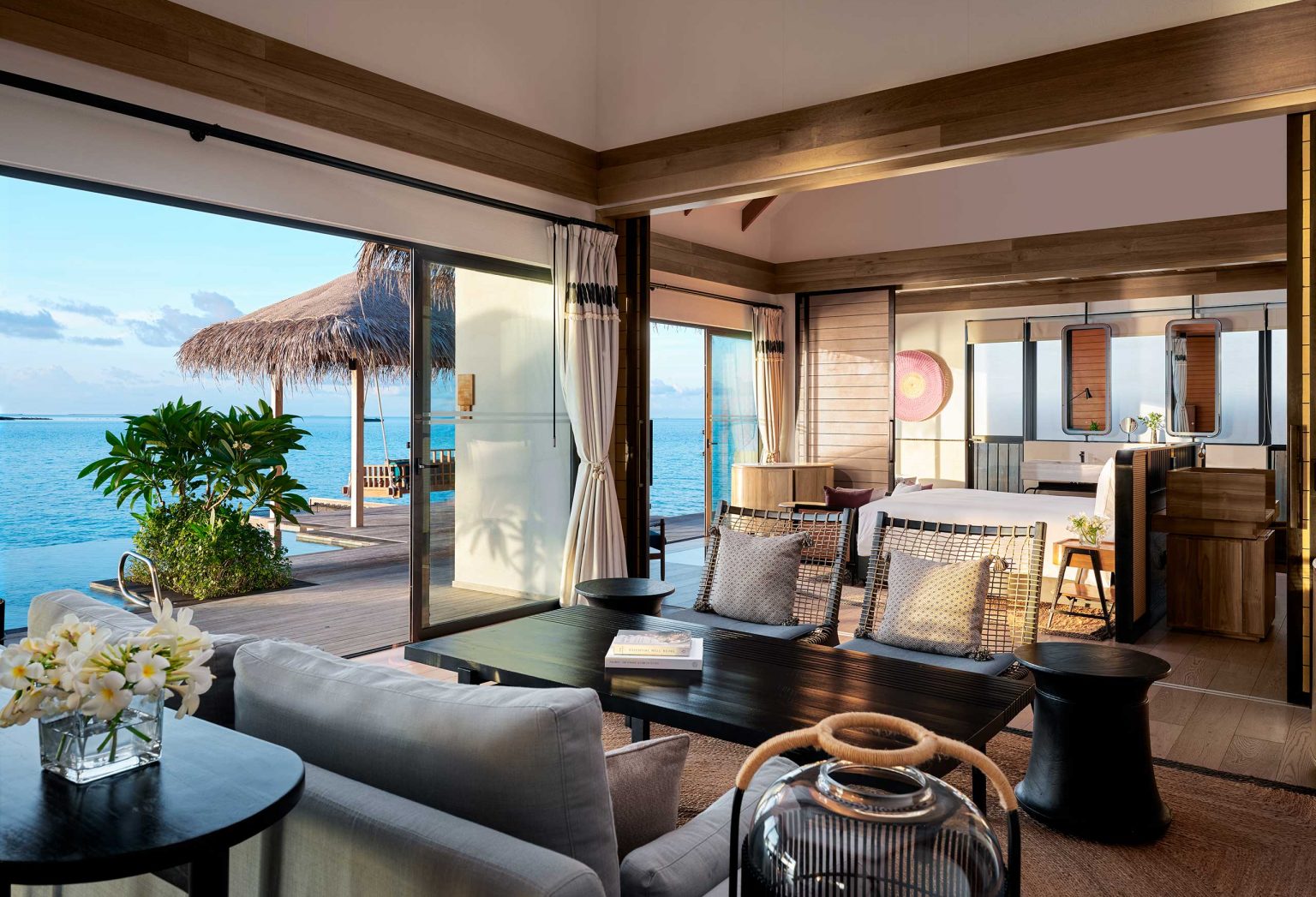 Luxurious open living space of a resort bungalow with light wood tones overlooks a serene ocean view. Large windows invite natural light. Cozy seating with tropical decor enhances tranquility.