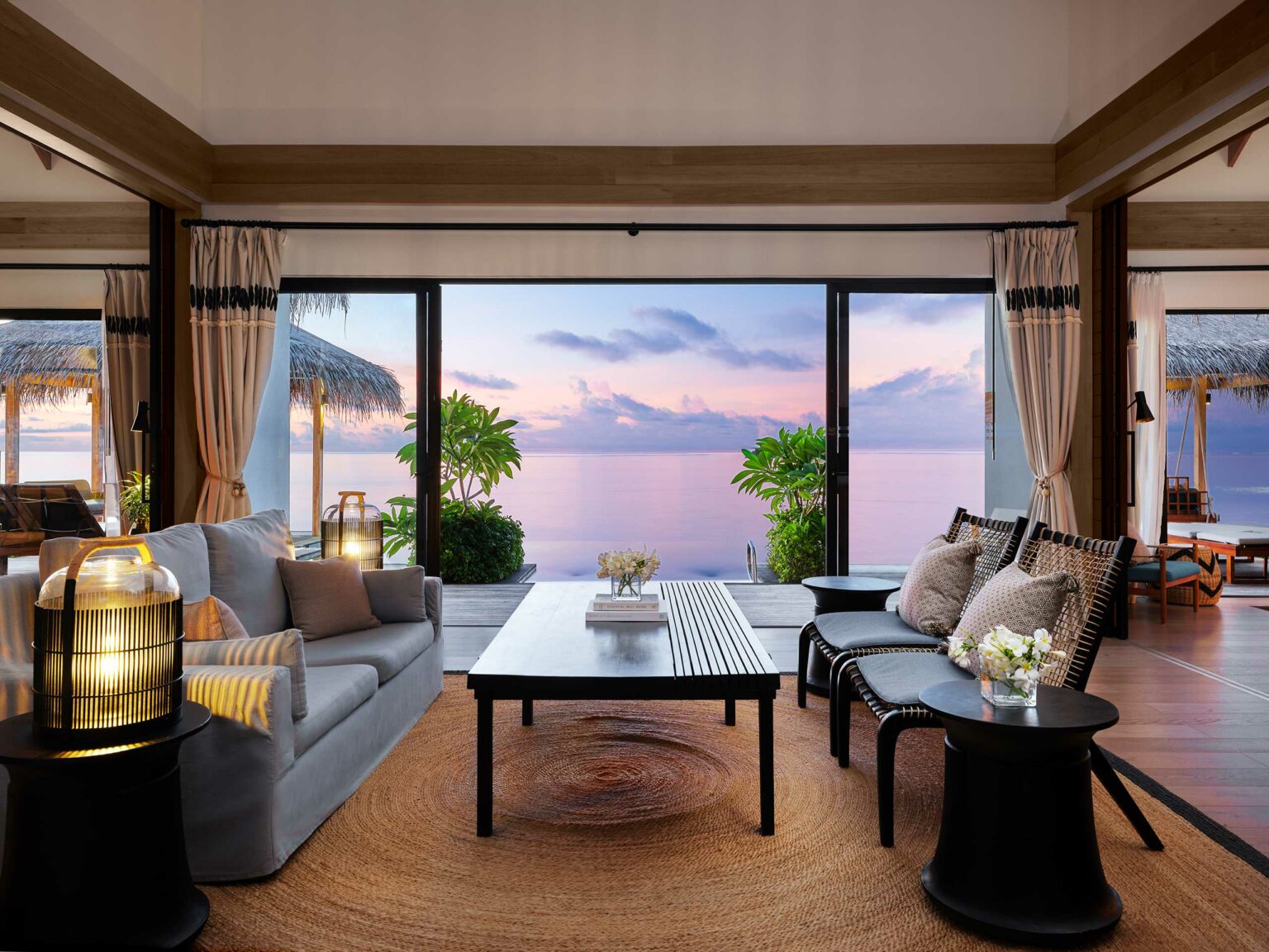 Elegant resort bungalow living room with a large window view of a serene ocean and purple sunset. Cozy sofas, lanterns, potted plants, and soft lighting create a tranquil ambiance.