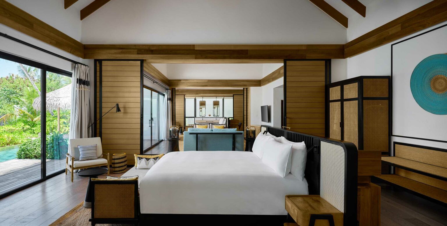 Spacious modern resort bedroom with wooden accents, large bed, blue sofa, and pool view via glass doors. Neutral tones create a relaxing ambiance.