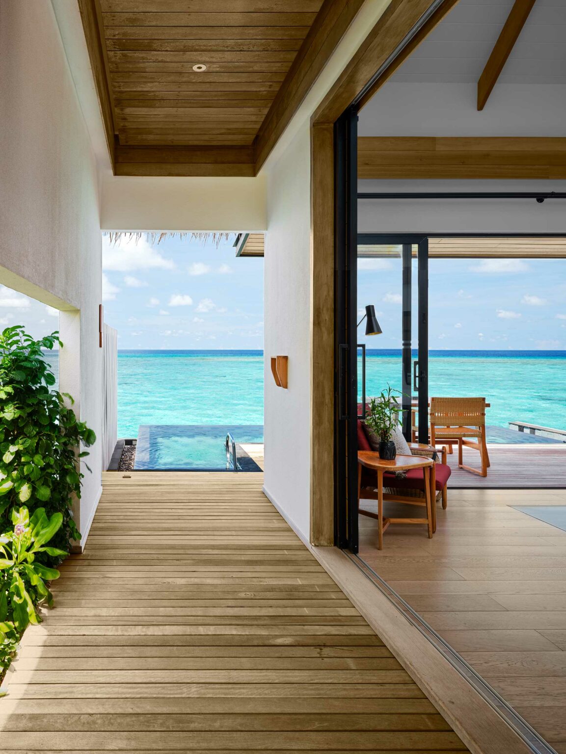 Luxurious resort beachfront bungalow with a wooden deck, infinity pool, and open living area. Overlooks tranquil turquoise ocean under a clear, sunny sky.