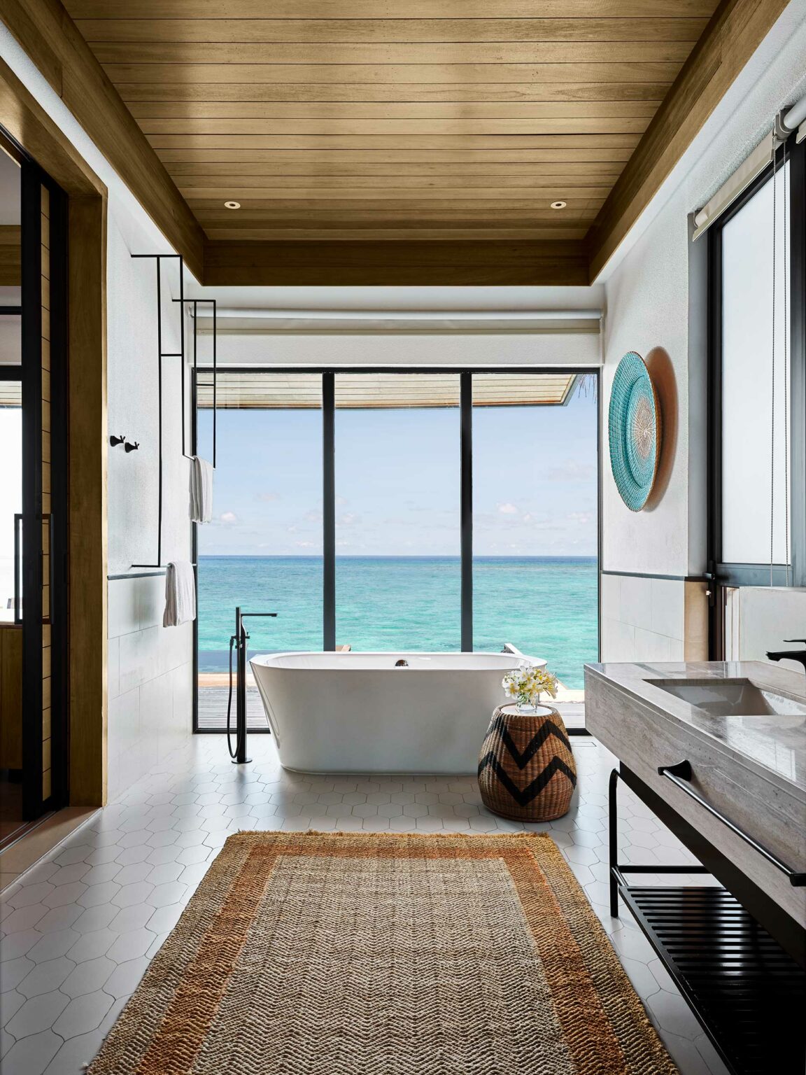 Luxurious suite bathroom with an ocean view through large windows. Features a freestanding bathtub, wooden ceiling, woven rug, and decorative vase, conveying tranquility.