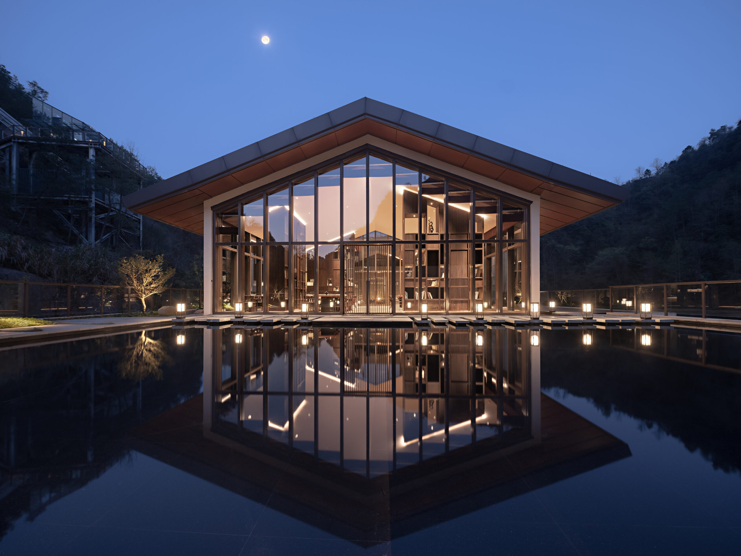 Modern glass pavilion at dusk, softly illuminated, reflects in a serene water pool. Surrounded by trees, it conveys elegance and tranquility.