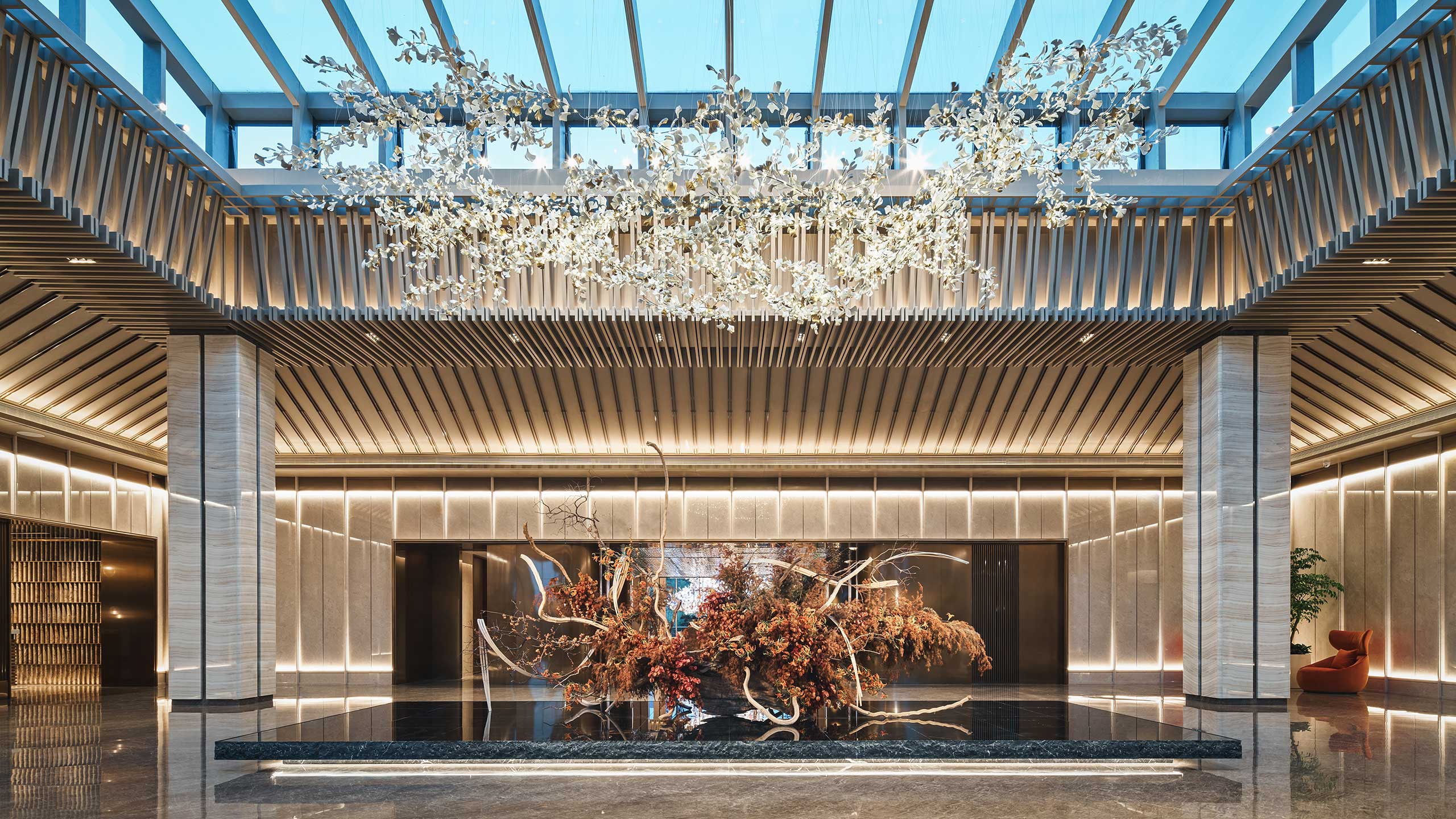 Luxurious modern hotel lobby with a sleek, reflective floor, large floral centerpiece, warm lighting, and an elaborate ceiling installation of delicate leaves.
