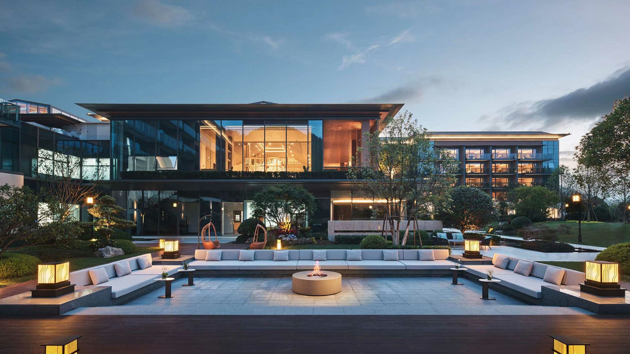 Modern hotel building with large glass windows illuminated at dusk. Foreground features a stylish courtyard with a round fire pit and elegant seating.