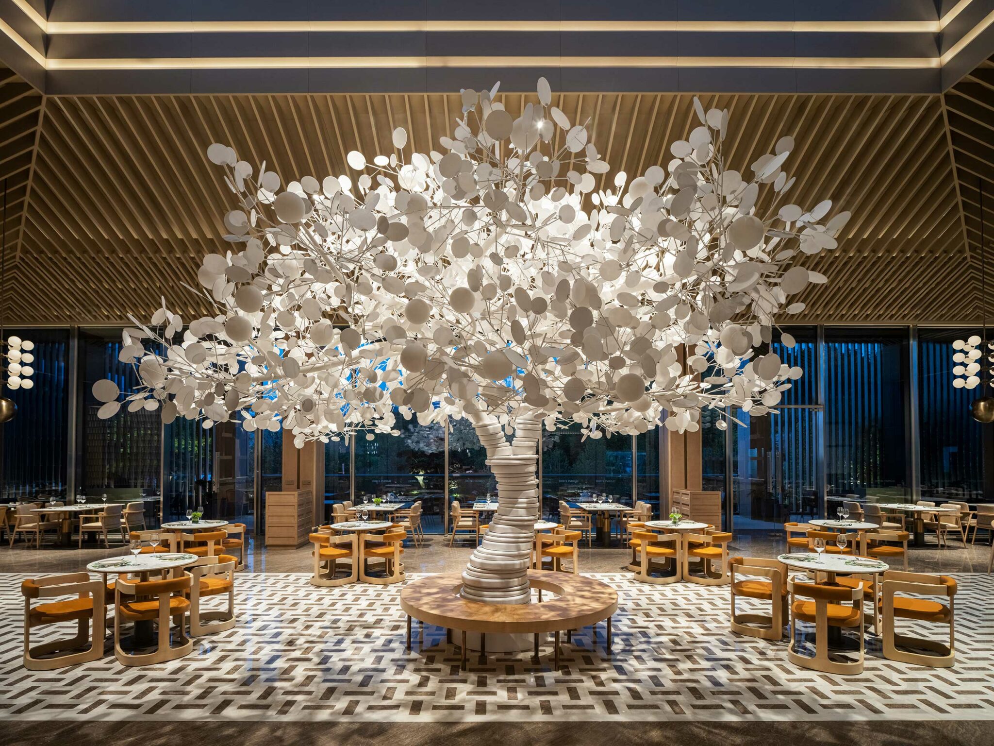 Modern hotel all day dining area with a striking white tree sculpture centerpiece featuring circular leaves, soft colored lights, and wooden seating. Elegant and creative.