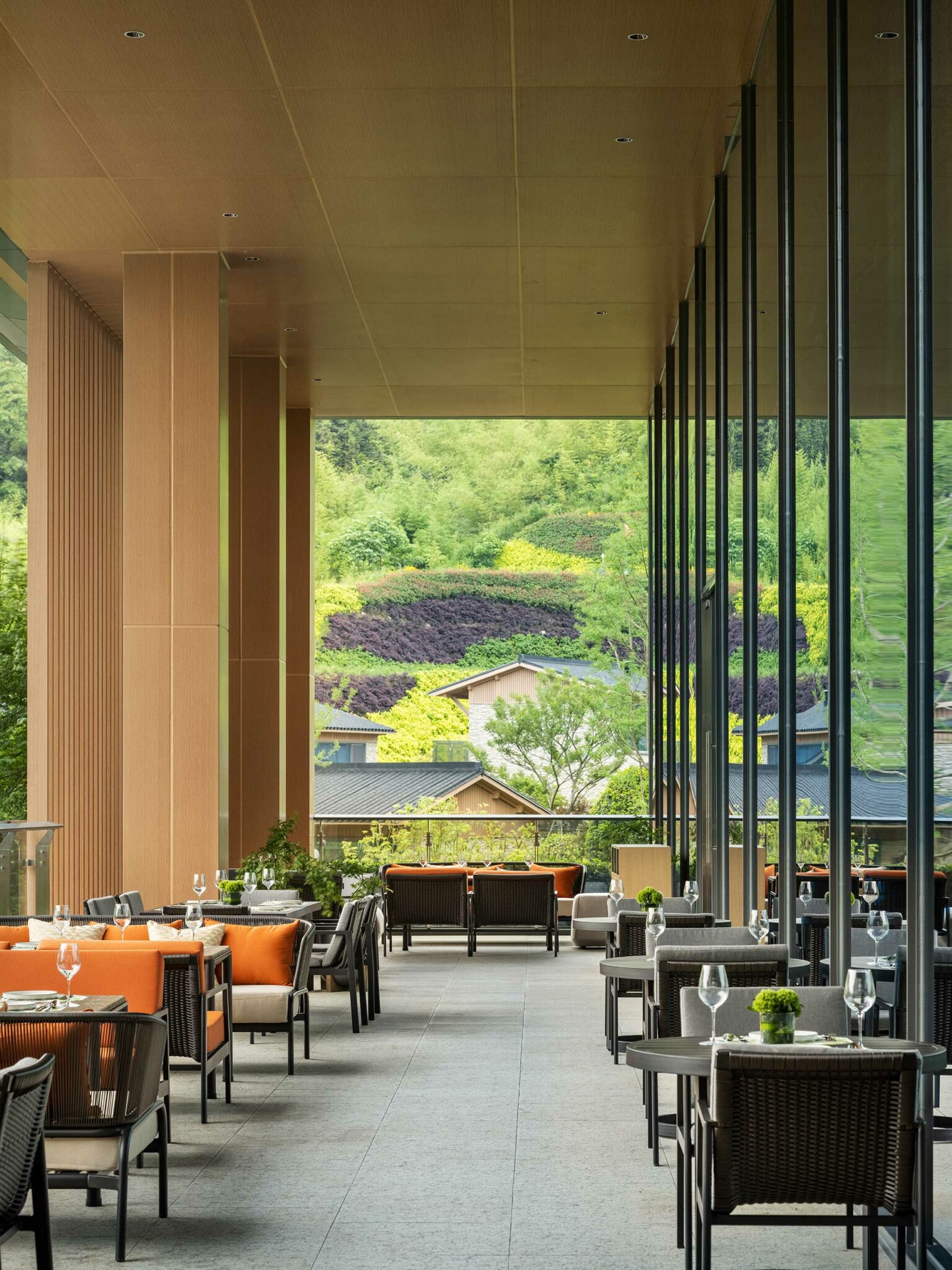 Stylish hotel outdoor dining area with wooden decor and glass walls overlooks a lush hillside with colorful plants. Tables are set with orange cushions.