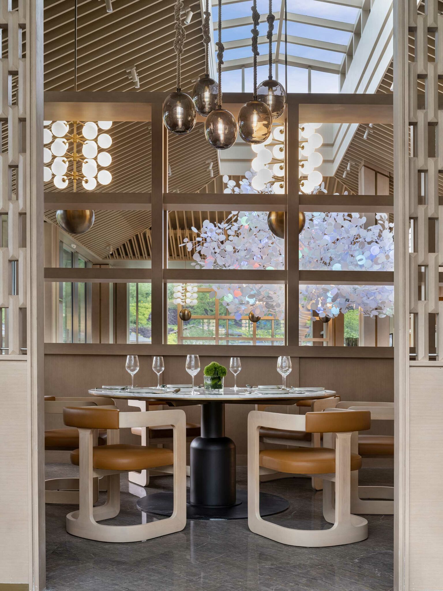 Elegant hotel dining area with a round table set for five, surrounded by modern wooden chairs. Overhead, artistic chandeliers and a lush outside view create a sophisticated ambiance.