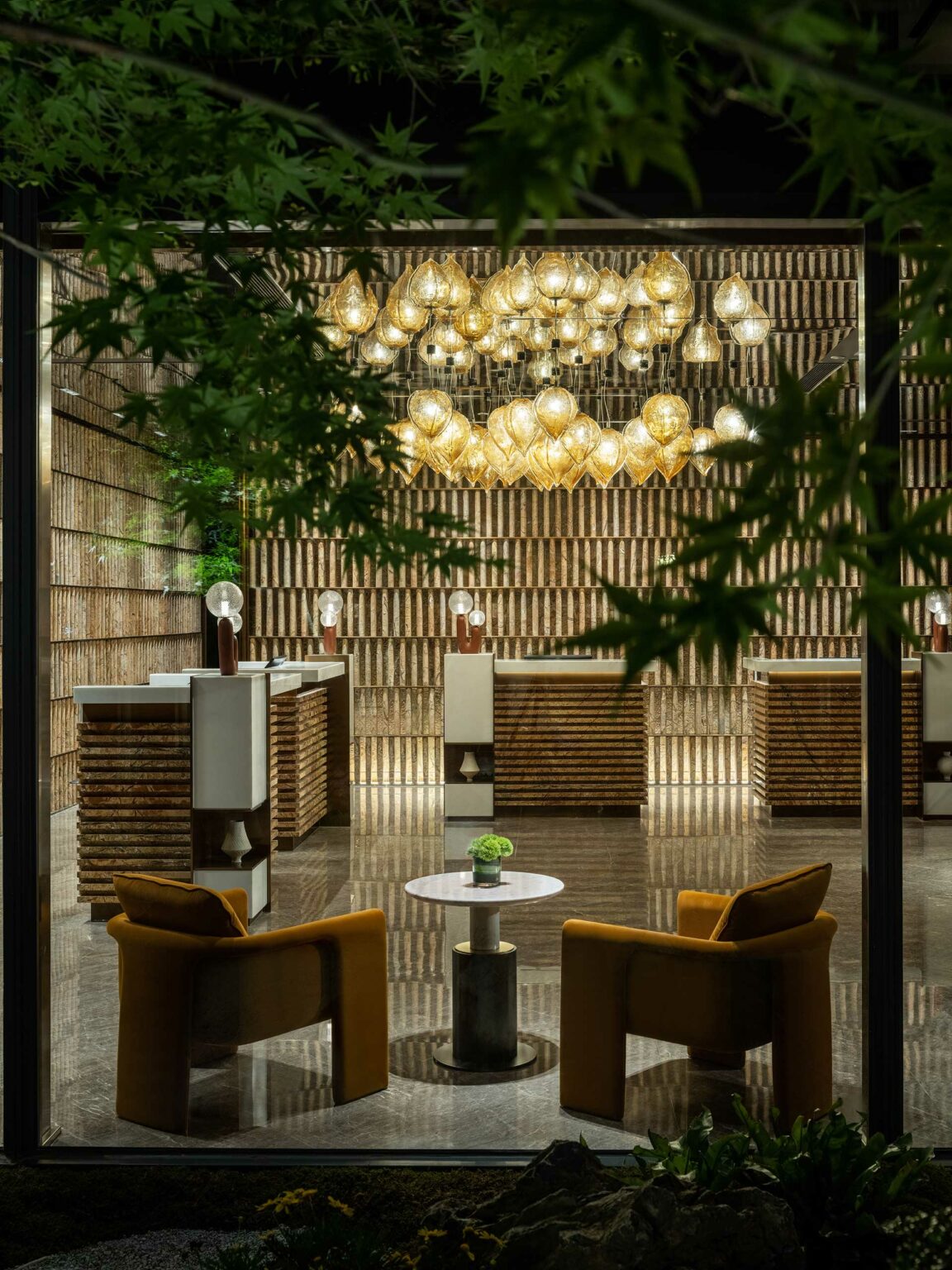 Luxurious hotel lobby with golden pendant lights, two mustard chairs, a small table, and a modern wooden reception desk. Green foliage frames the view.