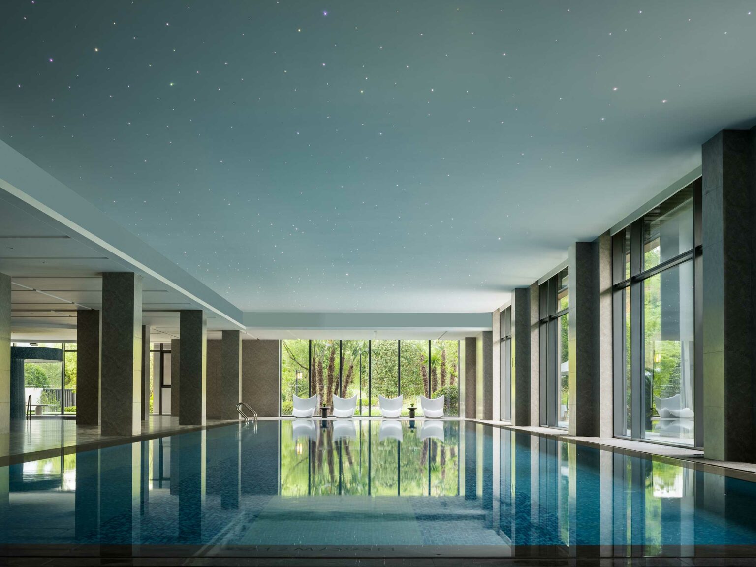 Spacious indoor hotel pool with reflective water, modern white lounge chairs, and large windows showcasing lush greenery. Ceiling resembles a starry night sky.