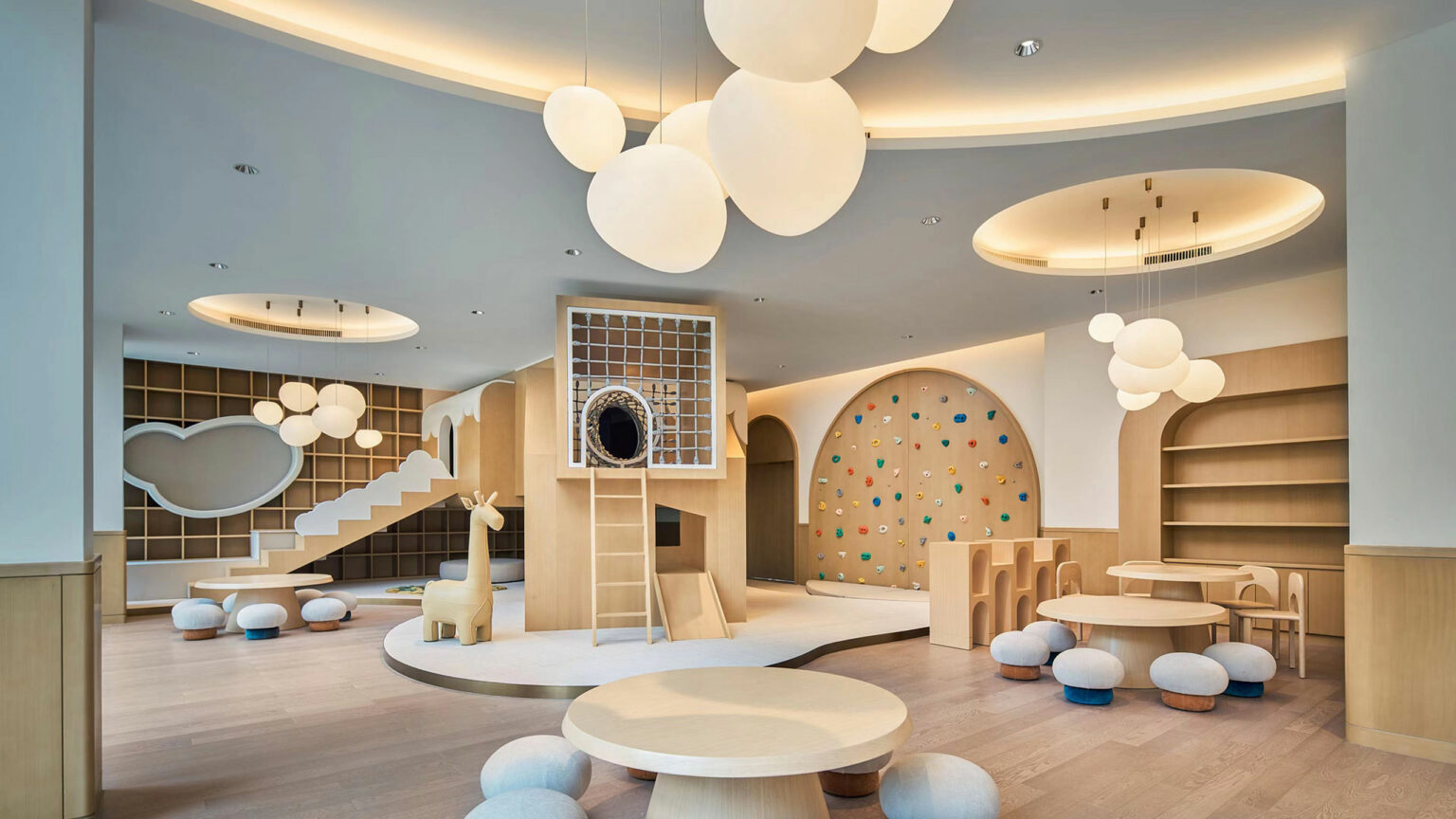 A spacious, modern hotel playroom with soft lighting features round tables, cushioned stools, a wooden giraffe, climbing wall, and whimsical ceiling lights for a playful, cozy atmosphere.
