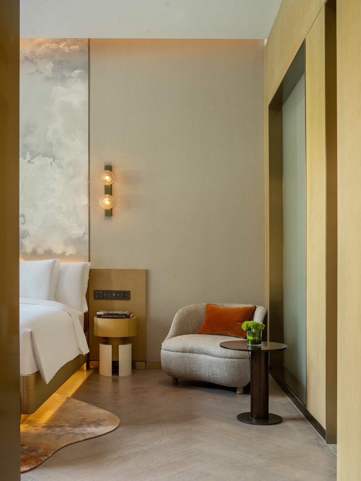A cozy hotel room corner with a cushioned armchair, orange pillow, and small side table holding a green plant. Beige walls add a warm, serene ambiance.