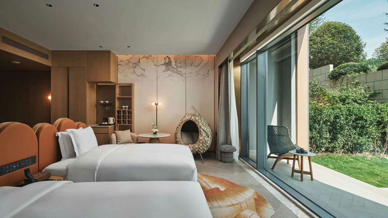 Modern hotel room with two white beds, orange headboards, and minimalist decor. Large window reveals a chair, table, and lush garden view outside. Cozy and serene atmosphere.