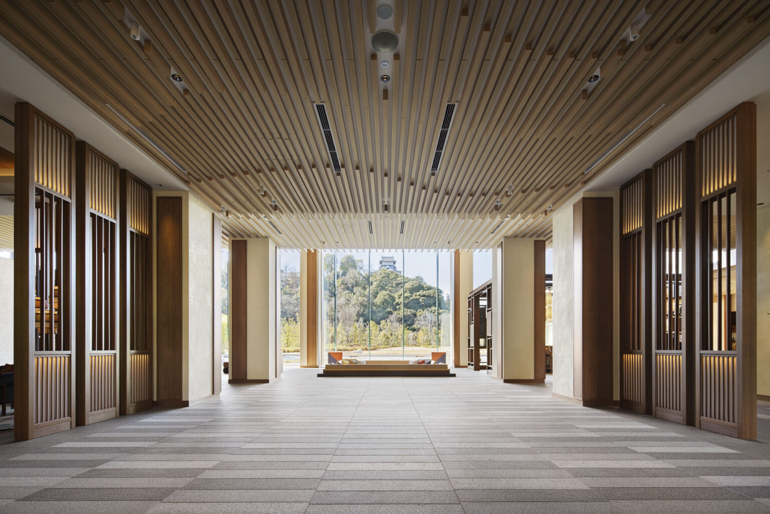 Spacious hotel hall with wooden slat ceiling and large windows showcasing a scenic view of trees and a hilltop building. Warm and inviting atmosphere.