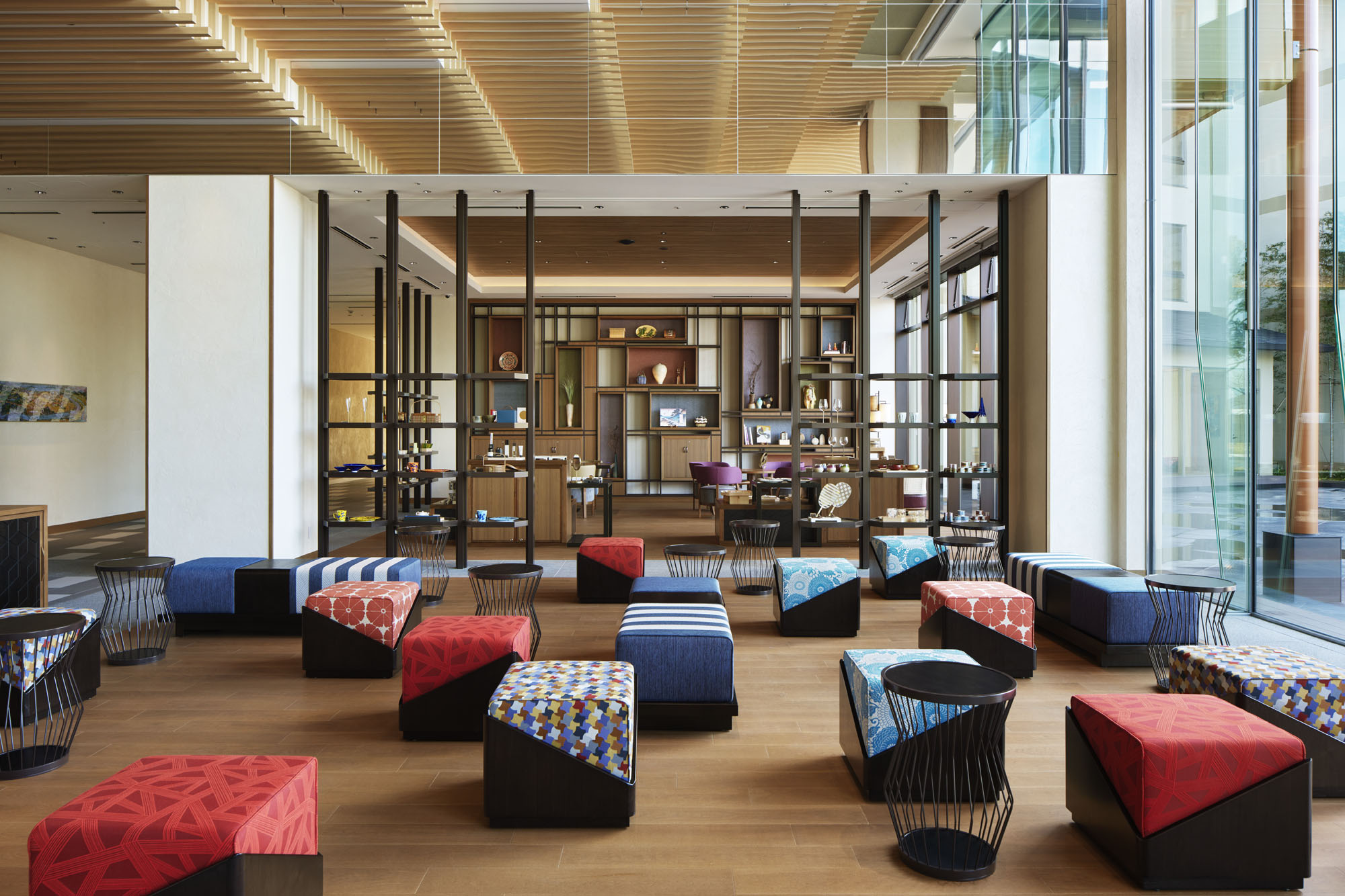 Modern hotel lounge area with colorful, patterned ottomans and small round tables on wooden floors. Large windows and bookshelves create an inviting atmosphere.