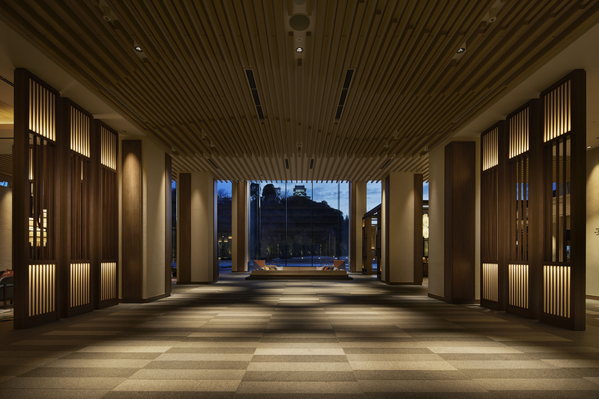 Spacious, elegant hotel hall with warm lighting, wooden accents, and large windows showcasing a serene evening view with distant trees and a structure.