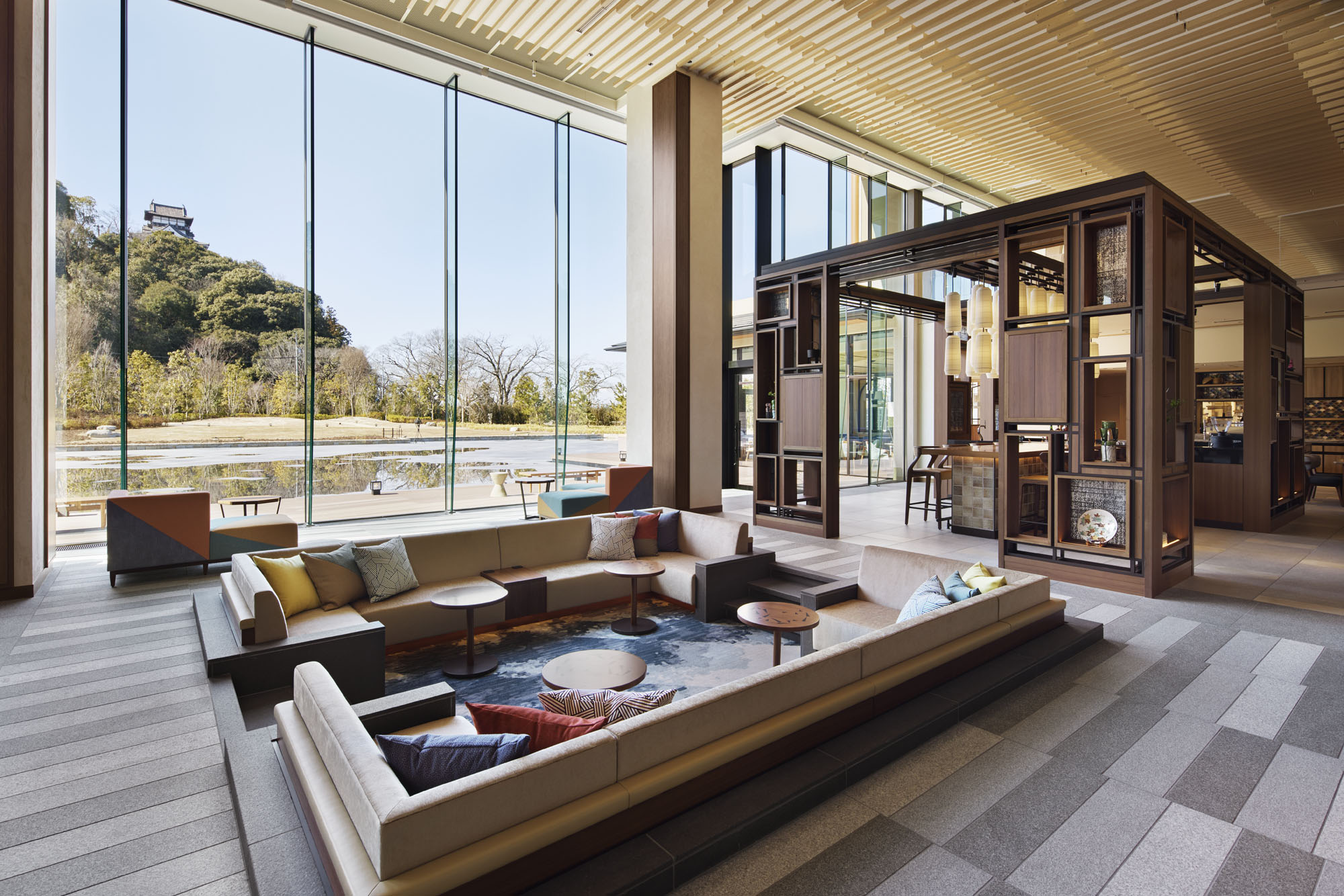 Modern hotel lounge with a sunken seating area, colorful cushions, and round tables. Large windows reveal scenic outdoor greenery and distant hills.